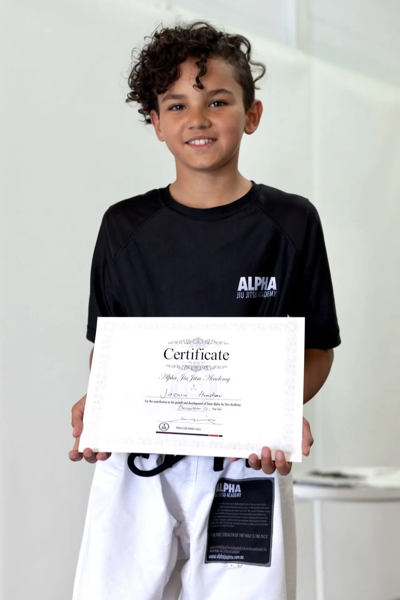 Jazmin Henshaw, a young student at Alpha Jiu-Jitsu Academy, proudly holds her graduation certificate while wearing a black academy shirt and white gi pants. She smiles confidently, showing her achievement within the program. The certificate features her name and signifies her dedication and progress. The simple background emphasizes Jazmin and her accomplishment at the academy.