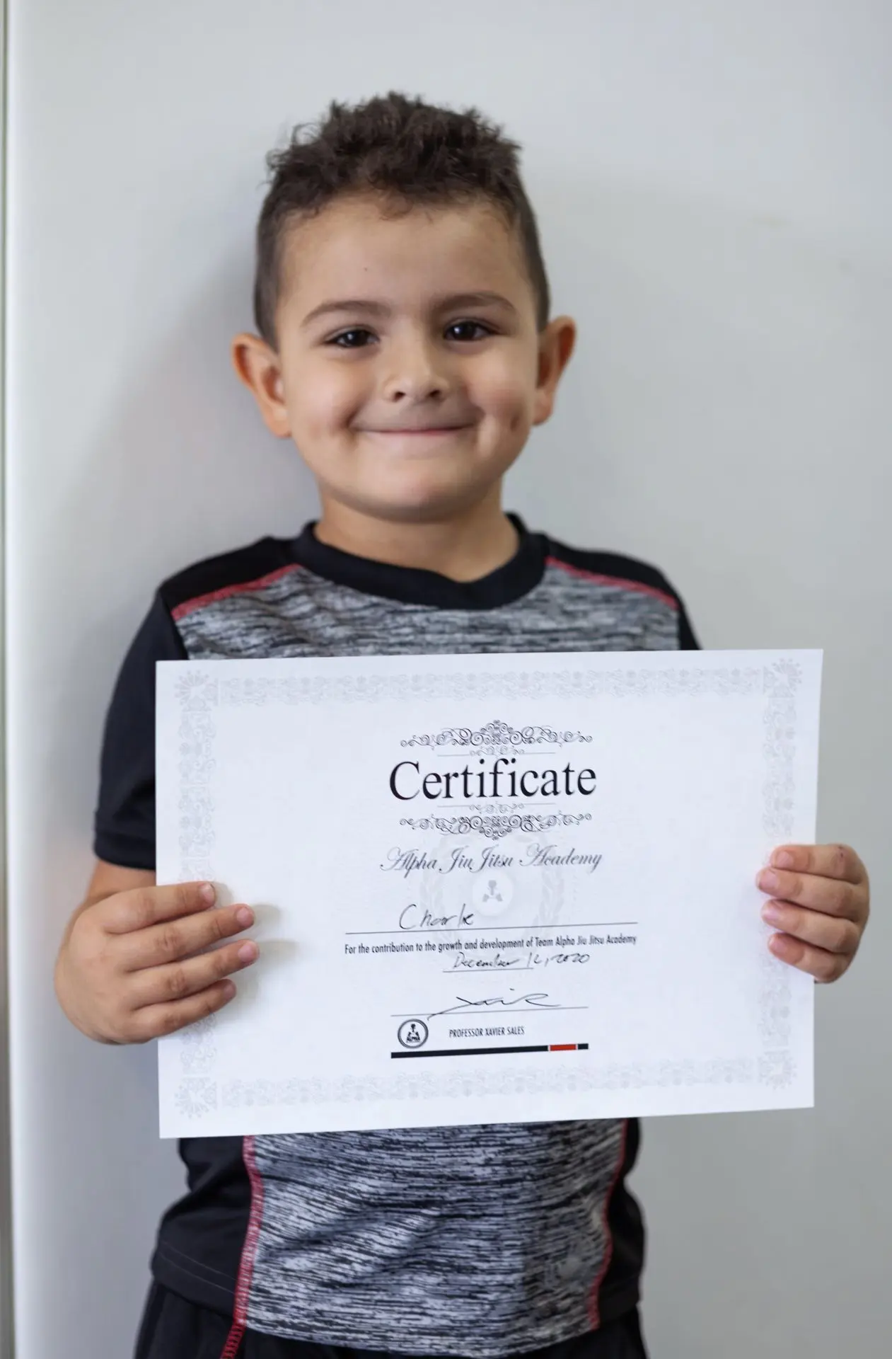 A young boy named Charlie proudly holding his certificate of achievement from Alpha Jiu-Jitsu Academy, smiling broadly with dimples and wearing a black and grey sports shirt. The certificate recognizes his dedication and contributions to his jiu-jitsu training, displayed against a neutral background.