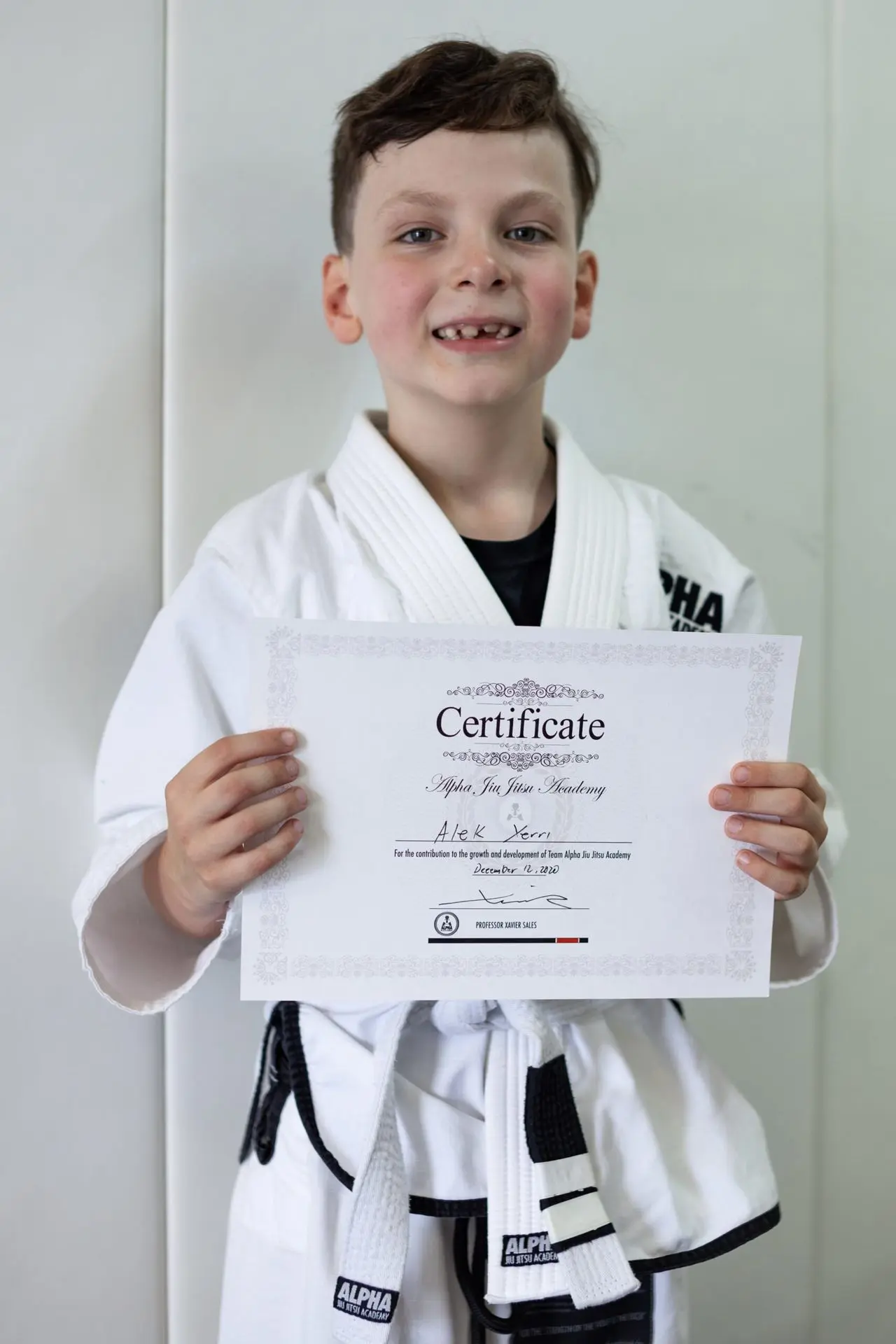 Alek Xerri, a young boy dressed in a white jiu-jitsu gi, holding his certificate of achievement from Alpha Jiu-Jitsu Academy, smiling proudly. The certificate acknowledges his growth and dedication to jiu-jitsu training, displayed against a plain background.