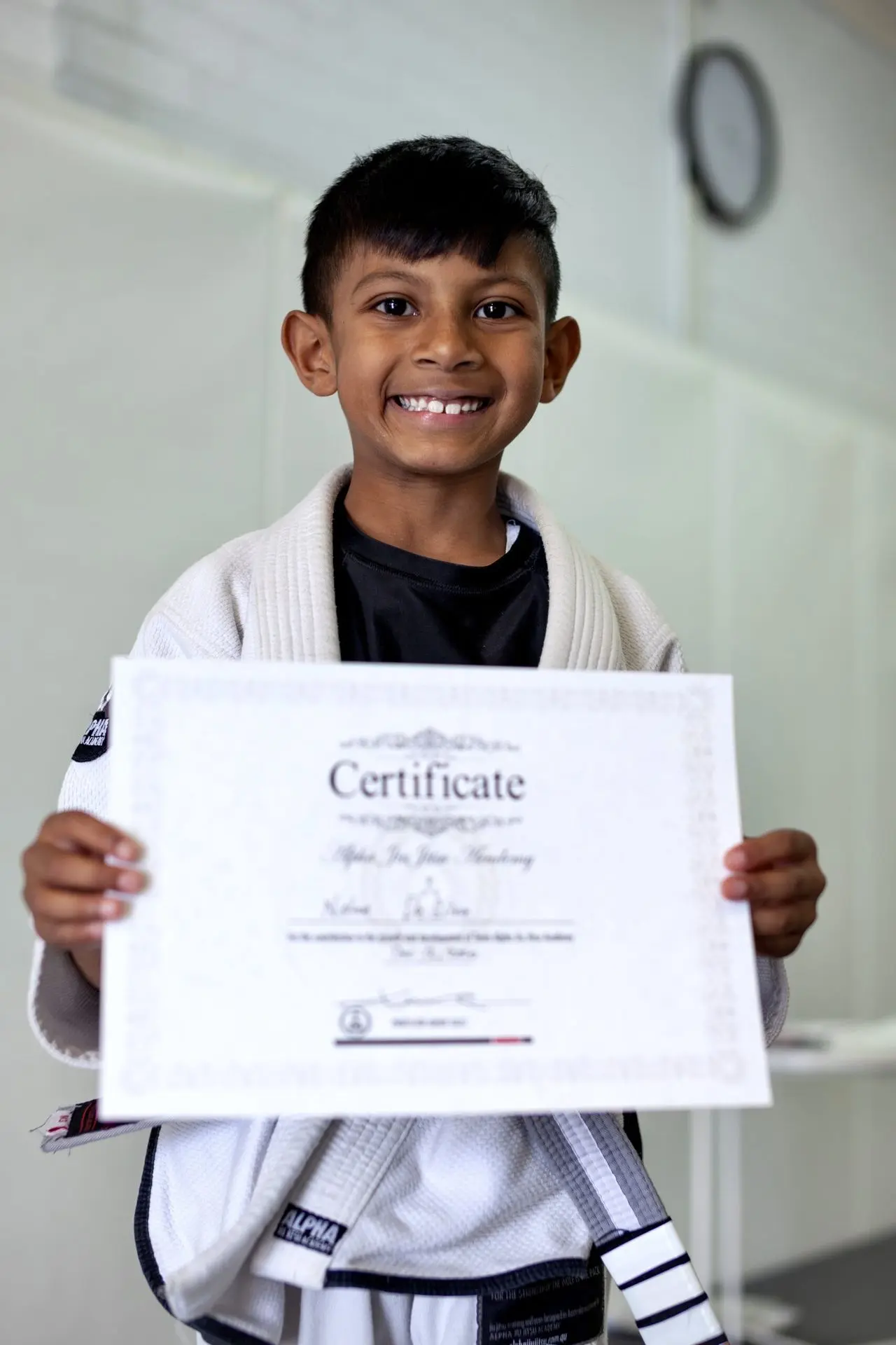 A young student at Alpha Jiu-Jitsu Academy smiling broadly while holding a certificate of achievement, wearing a white gi and standing proudly. The certificate marks a milestone in his training and dedication to jiu-jitsu. The background is plain, emphasizing the student's happy expression and accomplishment.