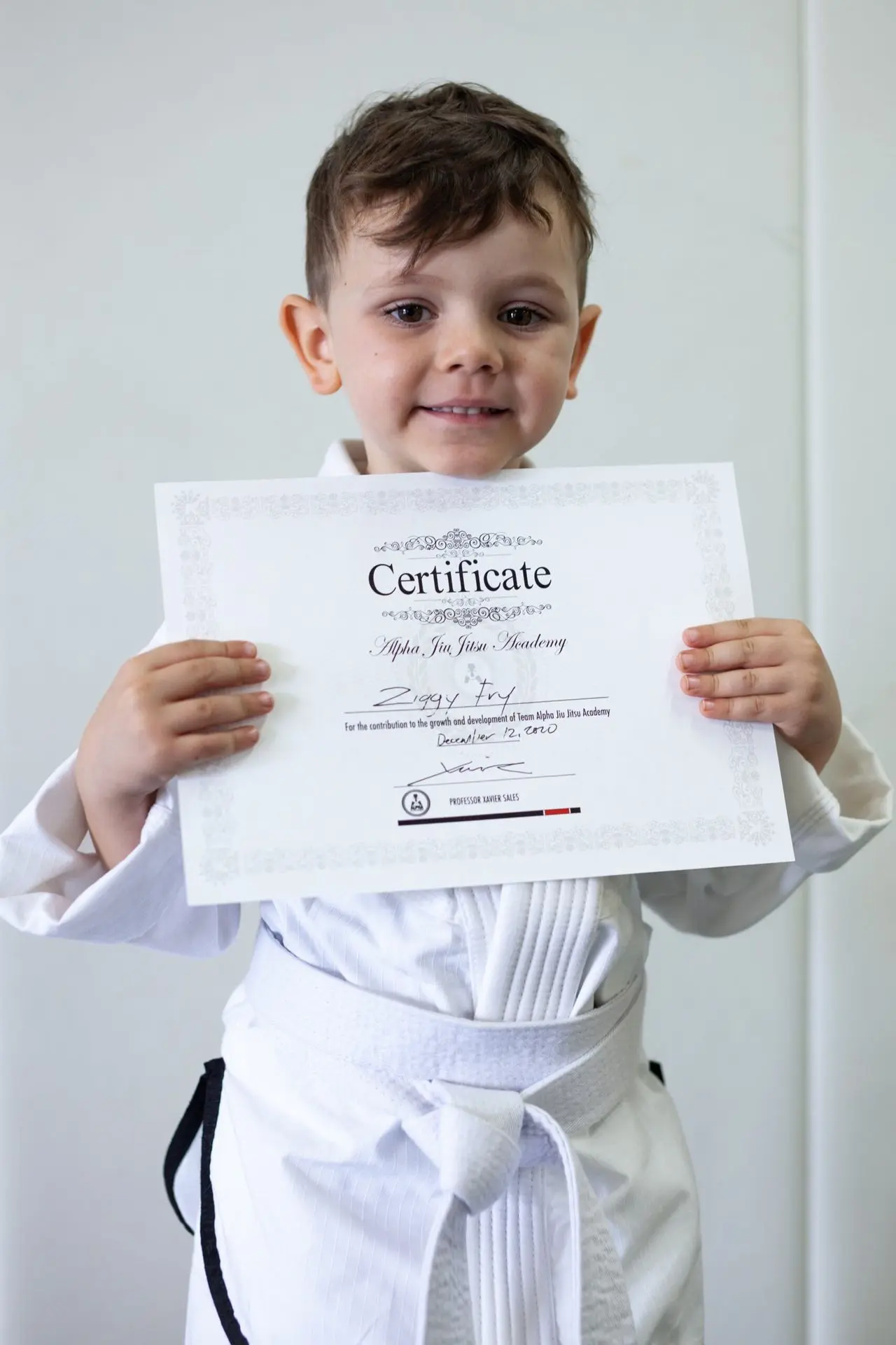 Ziggy Fry, a young boy in a white jiu-jitsu gi, proudly holding his certificate of achievement from Alpha Jiu-Jitsu Academy. The certificate acknowledges his growth and dedication to jiu-jitsu training. Ziggy is smiling against a plain background, showcasing his accomplishment.