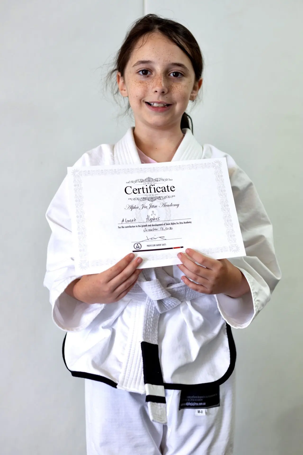 Hannah Hughes, a young girl wearing a white jiu-jitsu gi, proudly displays her certificate of achievement from Alpha Jiu-Jitsu Academy. She stands smiling and holding the certificate, which signifies her growth and dedication to her jiu-jitsu journey. The background is plain, highlighting her accomplishment.