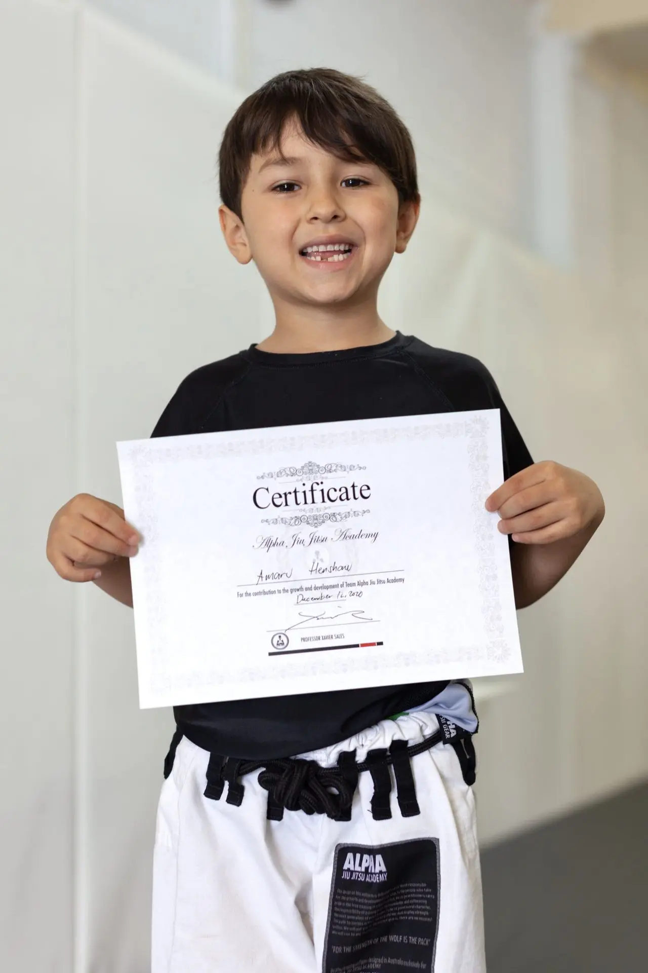 Amaru Henshaw, a young student at Alpha Jiu-Jitsu Academy, proudly holds his graduation certificate while wearing a black top and white gi pants. He smiles widely, celebrating his achievement. The certificate, displaying his name, symbolizes his progress and dedication within the academy's program. The clean background emphasizes Amaru and his milestone accomplishment.