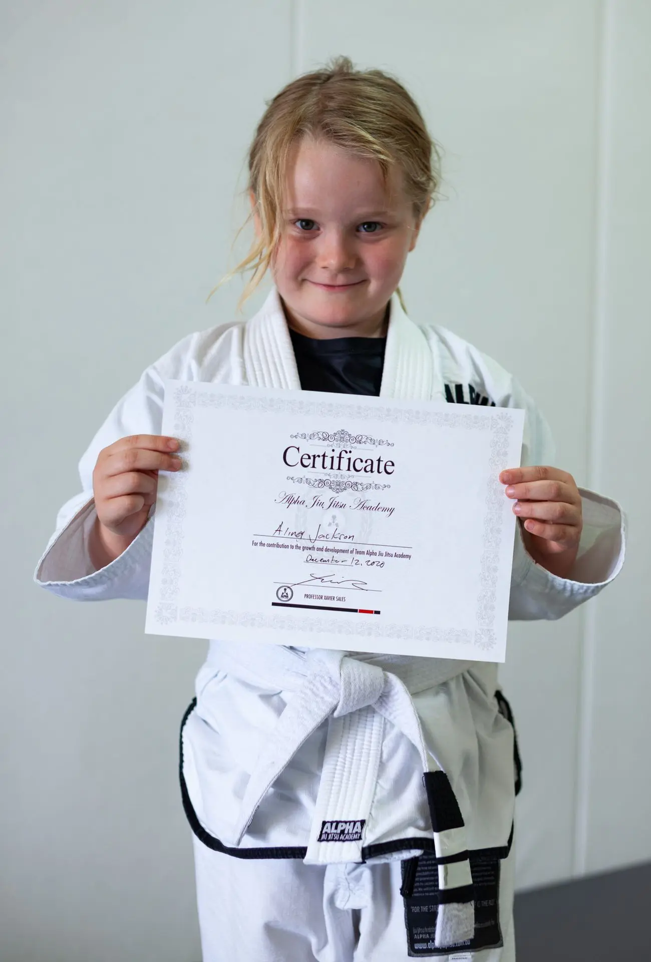 Aimey Jackson, a young girl in a white jiu-jitsu gi, smiles while proudly holding her certificate of achievement from Alpha Jiu-Jitsu Academy. Her hair is lightly tied back, and she is standing against a plain backdrop, highlighting her accomplishment and dedication.