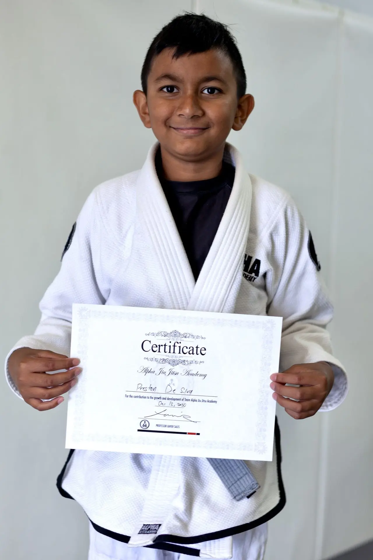 Preston De Silva, a young boy wearing a white jiu-jitsu gi, proudly holds his certificate of achievement from Alpha Jiu-Jitsu Academy. He smiles confidently, showcasing his dedication and accomplishment. The background is simple, putting emphasis on Preston and his certificate.