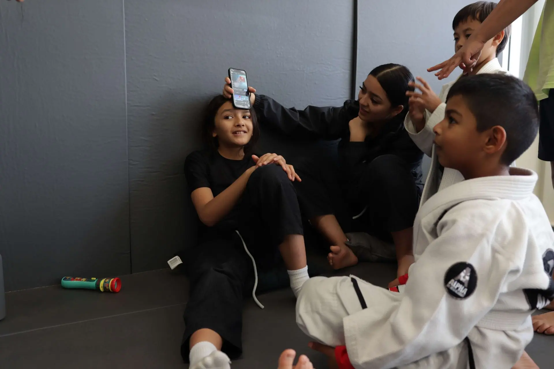 A group of children socializing after their Brazilian Jiu-Jitsu class at Alpha Jiu-Jitsu Academy. One child sits on the mat, smiling while a phone is held above their head. The other children around her are engaged, with one child holding the phone and others interacting in the background. Some kids are still in their gis, while others wear athletic clothing. The atmosphere is playful and light-hearted, capturing the fun, post-training camaraderie among the students.