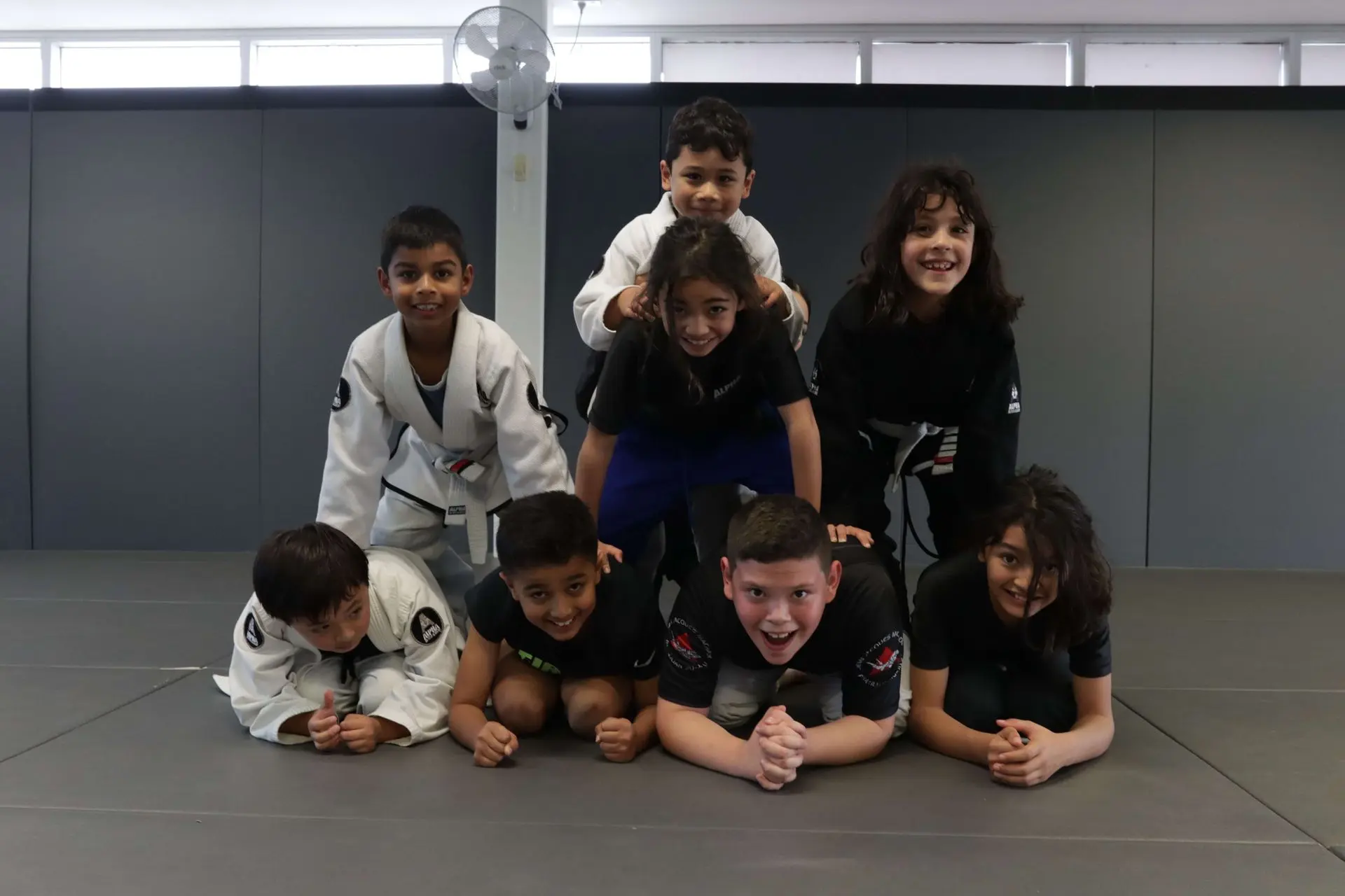 A playful group of children is posing in a human pyramid formation at Alpha Jiu-Jitsu Academy after their Jiu-Jitsu class. The kids are all smiling, some in gi uniforms, while others wear athletic gear. They seem to be having fun as they show off their teamwork and friendship. The photo captures a lively, cheerful moment, with the academy's training area in the background, showcasing the camaraderie and positive atmosphere among the students.