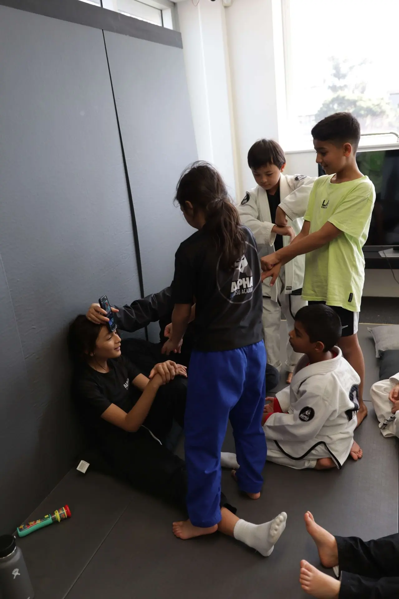 A group of children gathered after a Brazilian Jiu-Jitsu session at Alpha Jiu-Jitsu Academy. Some are sitting on the mats, while others are playfully interacting. One child is sitting with a cell phone held to another child's forehead, possibly taking a photo. In the background, other students in gi and athletic clothing stand and engage with their peers. The academy's logo is visible on their gear. The room has a relaxed, social atmosphere with students laughing and bonding.