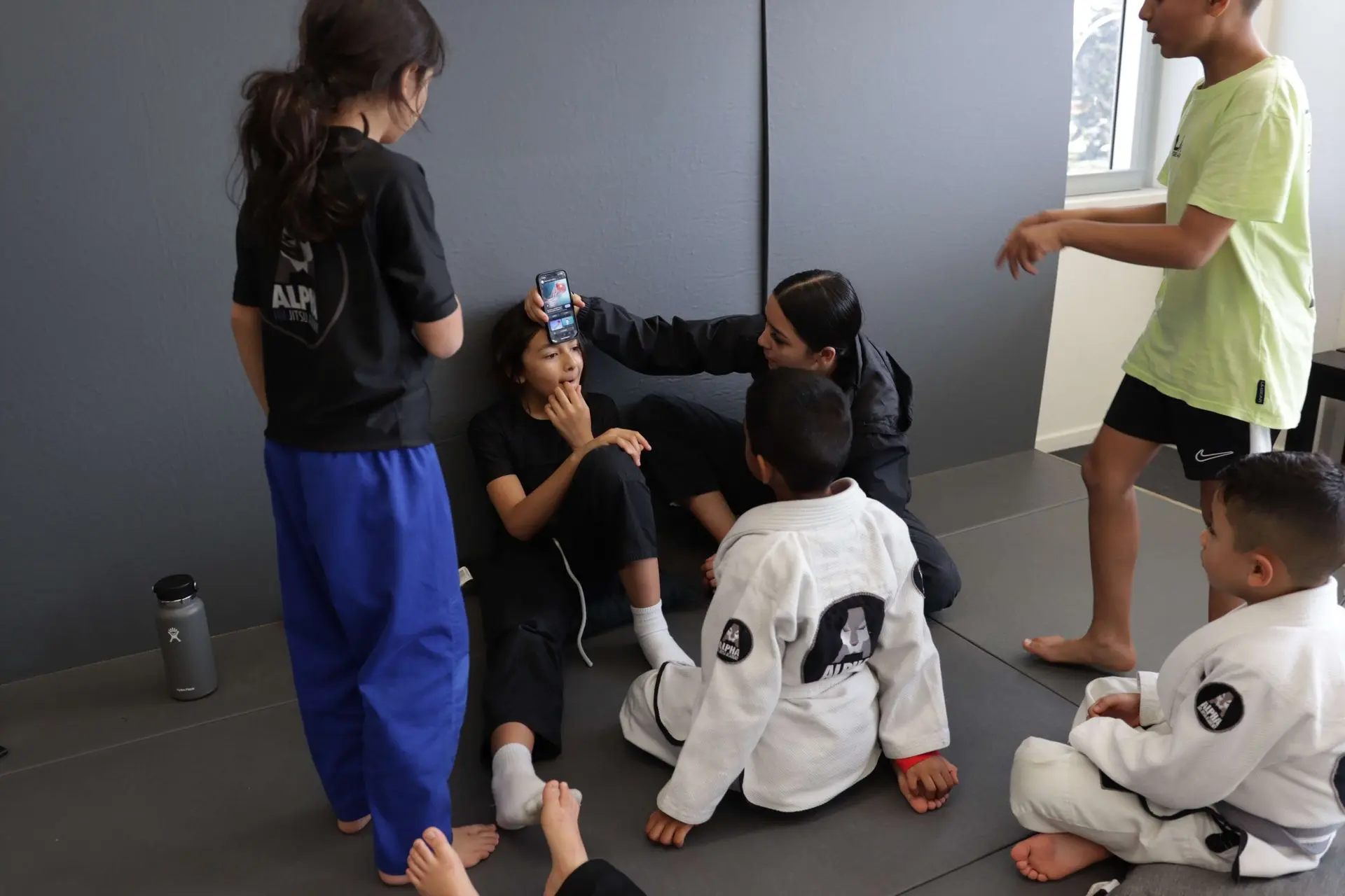 The image shows a group of Alpha Jiu-Jitsu Academy students taking a break during a training session. A female instructor is using a phone to monitor a student's temperature or other activity as part of a fun exercise, while other students interact playfully around her. The children are dressed in Alpha Jiu-Jitsu branded shirts and gis, with one child in a green gi and another in a yellow t-shirt. Several students are sitting on the mat, engaging in conversation or waiting for instructions, while others stand nearby. The atmosphere is relaxed and friendly, with the academy's gray-colored training mats and modern interior in the background.