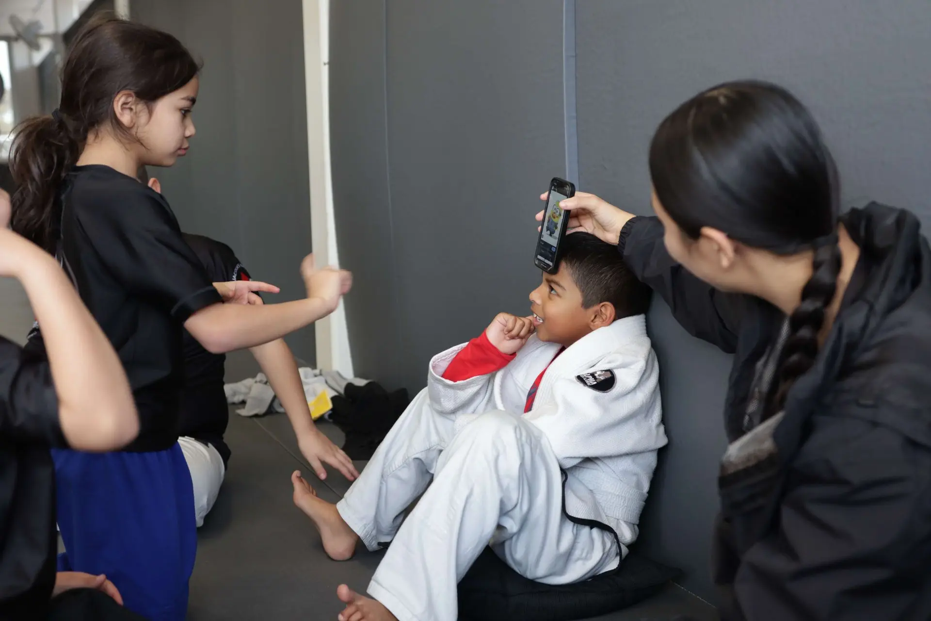 The image shows a group of young students from Alpha Jiu-Jitsu Academy engaged in a fun and educational moment. A female instructor is using a phone to show something to a student, who is seated and intently looking at the screen. Another student in a black shirt is interacting nearby, while the others are seated or standing around, actively participating in the moment. The students are dressed in Alpha Jiu-Jitsu branded gear, with some in gis and others in academy t-shirts. The atmosphere appears relaxed and focused, with a lighthearted interaction taking place in a modern training space.