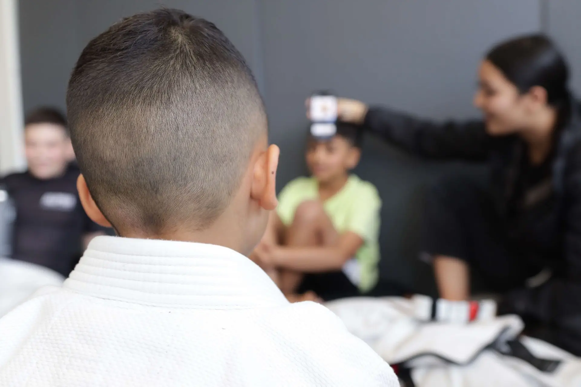 The back of a child's head in a white gi is in the foreground, with a few other children visible in the background. One child is sitting with their arms raised, while another is smiling, as an adult in black clothing appears to be interacting with them. The focus is on the child in the foreground, while the others are slightly blurred.