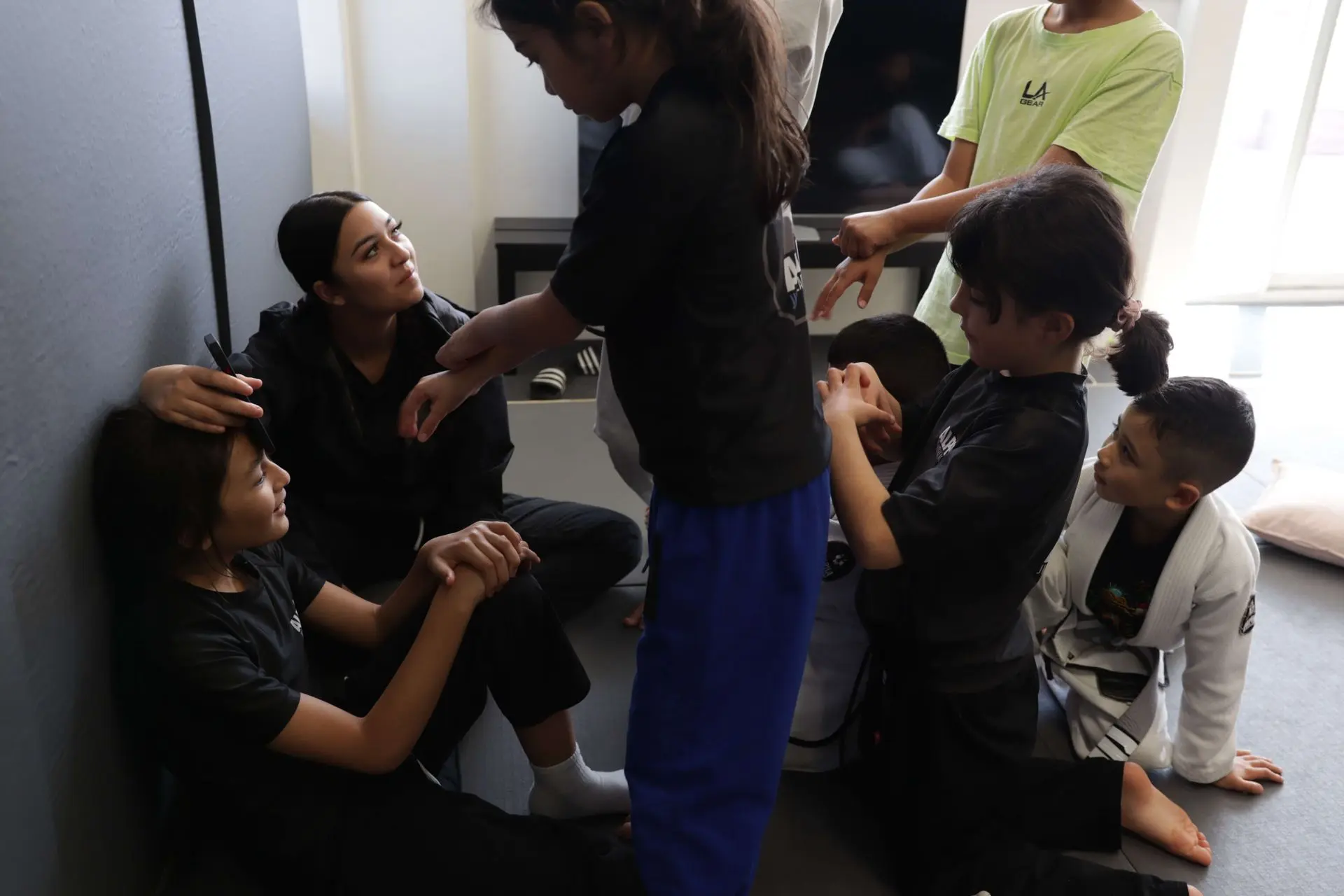 A group of children is engaged in fun post-class interactions at Alpha Jiu-Jitsu Academy. In the center, a child sits on the mat, smiling as another child holds a phone over her head. A few other kids are gathered around, interacting playfully, while a coach or older student watches and smiles. The children are in casual athletic clothing, some still in their gi, and the atmosphere is light-hearted and cheerful, showcasing the camaraderie among the students.