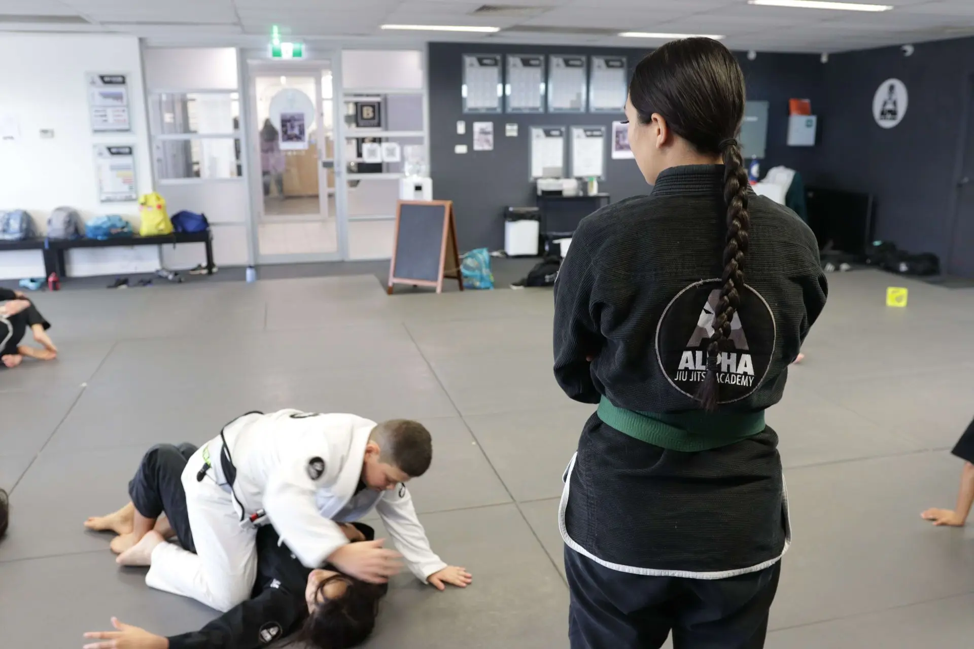 In this image, a Brazilian Jiu-Jitsu instructor wearing a black gi with a green belt is standing at the edge of the mat, observing students during a practice session at Alpha Jiu-Jitsu Academy. A student in a white gi is shown performing a top position drill on a classmate wearing a black gi. In the background, other students are engaged in different grappling techniques. The academy’s walls feature certificates and a logo, and the space is well-lit with a welcoming atmosphere. The instructor is focused on providing guidance, watching over the students' progress.