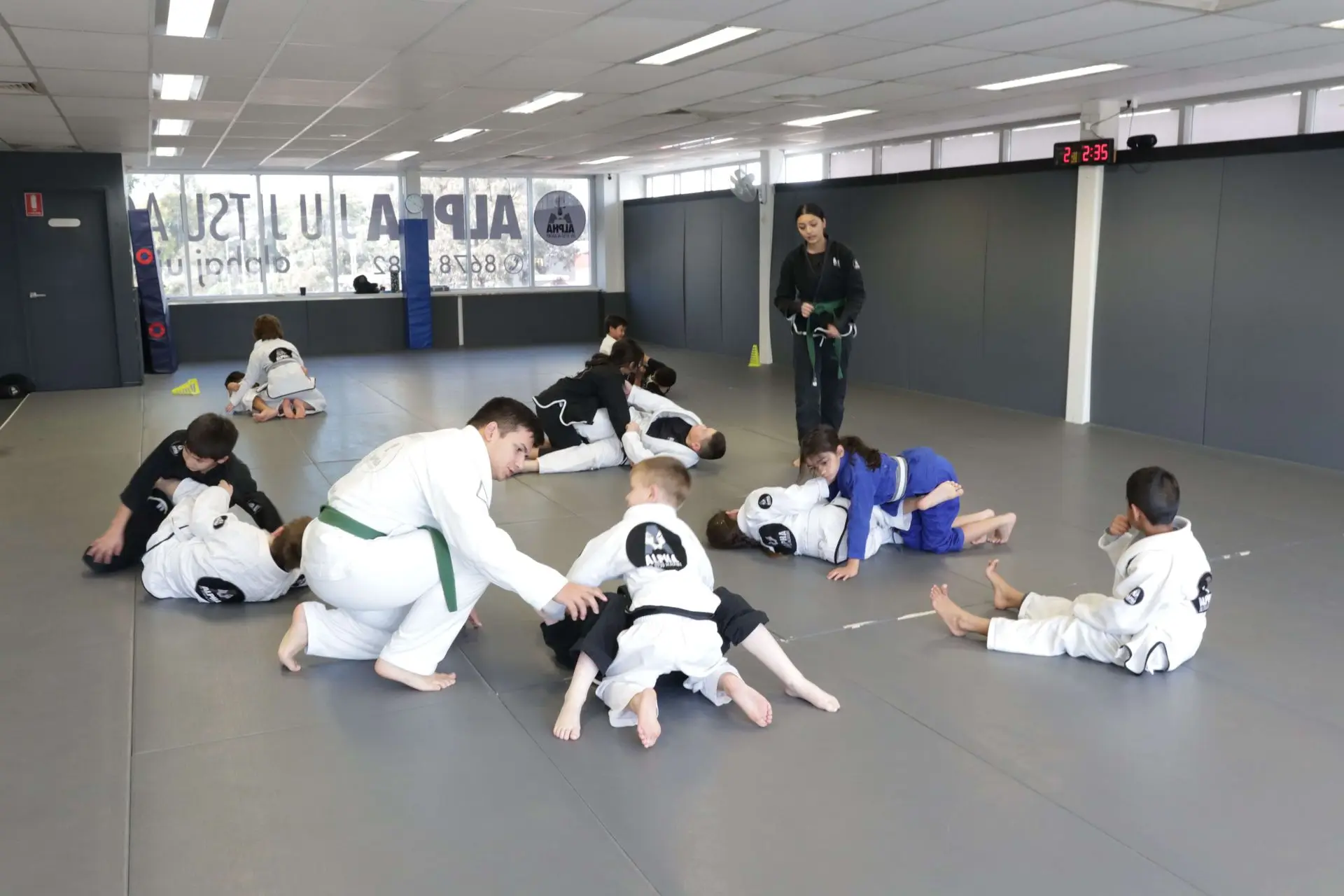 The image depicts a Brazilian Jiu-Jitsu class in action at Alpha Jiu-Jitsu Academy. A green belt instructor is guiding students as they practice different grappling techniques. Some students in white gis are rolling with each other on the mat while others, wearing black and blue gis, are positioned in various positions, refining their skills. The instructor observes and offers guidance, while a large clock displays the remaining class time. The academy’s spacious, well-lit training area features the Alpha Jiu-Jitsu logo on the walls.