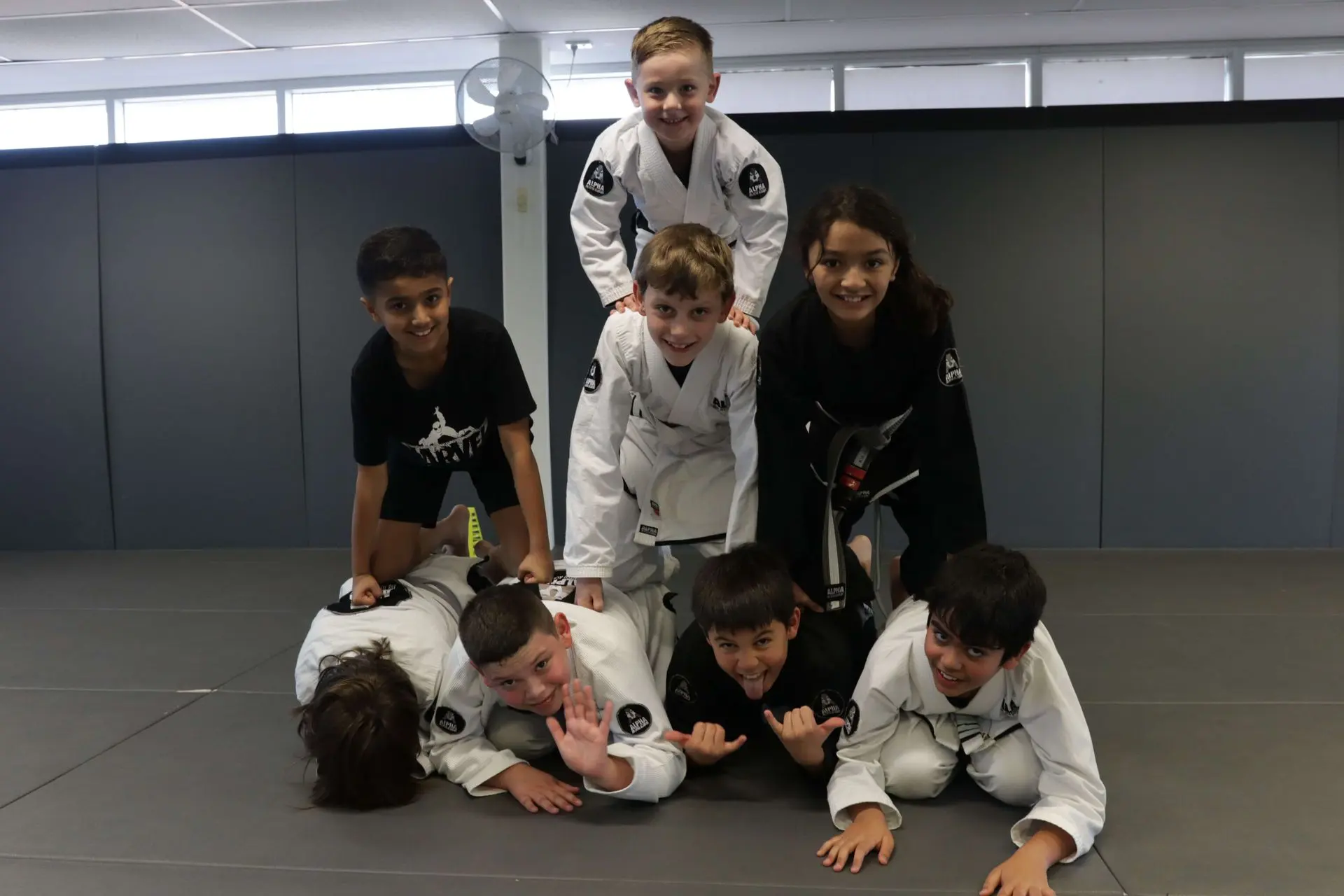 A group of seven kids from Alpha Jiu-Jitsu Academy are playfully stacked in a human pyramid on the mat. Some of the children are wearing white gi with the academy's logo, while others are in black gi. They are all smiling and laughing, enjoying the fun moment. The background shows the academy’s training space with a fan on the wall and bright natural light.