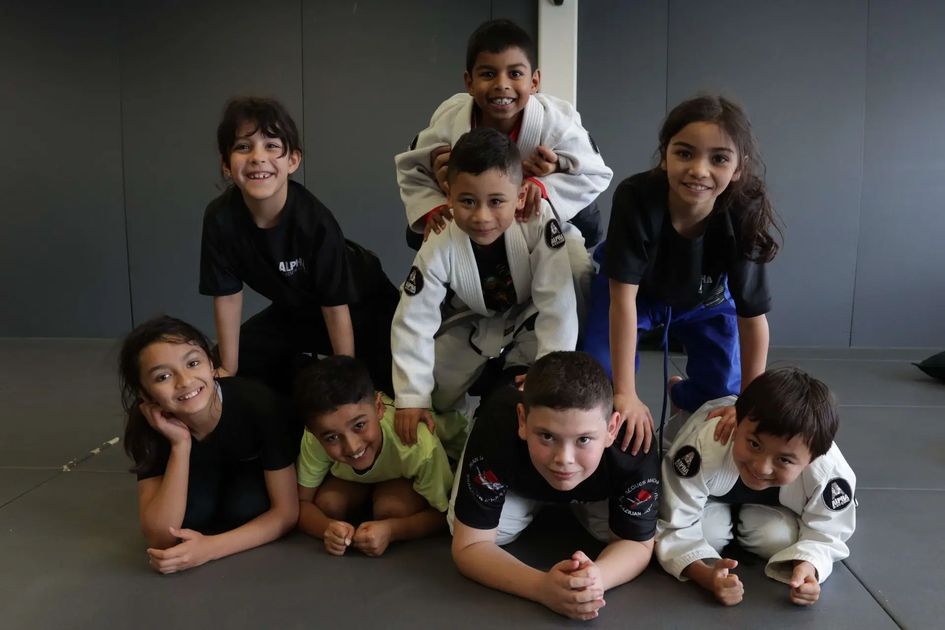 A group of eight children, both boys and girls, are smiling and posing while forming a human pyramid on the mat at Alpha Jiu-Jitsu Academy. They are dressed in either black t-shirts or gi uniforms. The child on top, wearing a gi, smiles as they pose for the photo, while the others underneath them are either kneeling or lying on the floor, grinning. The background features a plain gray wall and the academy’s mat area, adding to the joyful atmosphere of the playful scene.