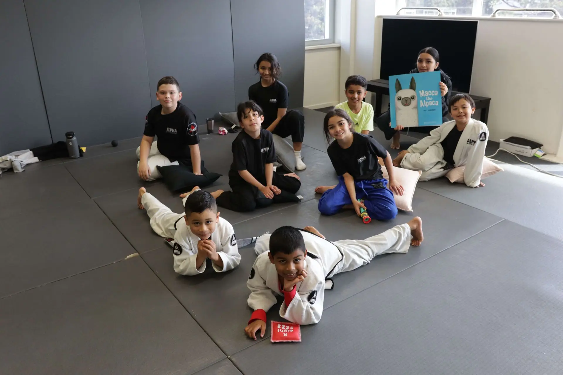 A group of children is gathered on the mat at Alpha Jiu-Jitsu Academy. Some of them are dressed in black academy t-shirts, while others are in gi uniforms. They are posing for a photo, with a girl holding a picture book titled "Macca the Alpaca" in front of her. A boy is lying on the floor in the foreground, resting his head on his hands, while another boy and girl are sitting with their arms around each other. In the background, a few other children are lounging and engaging in light conversation. The atmosphere is fun and relaxed.