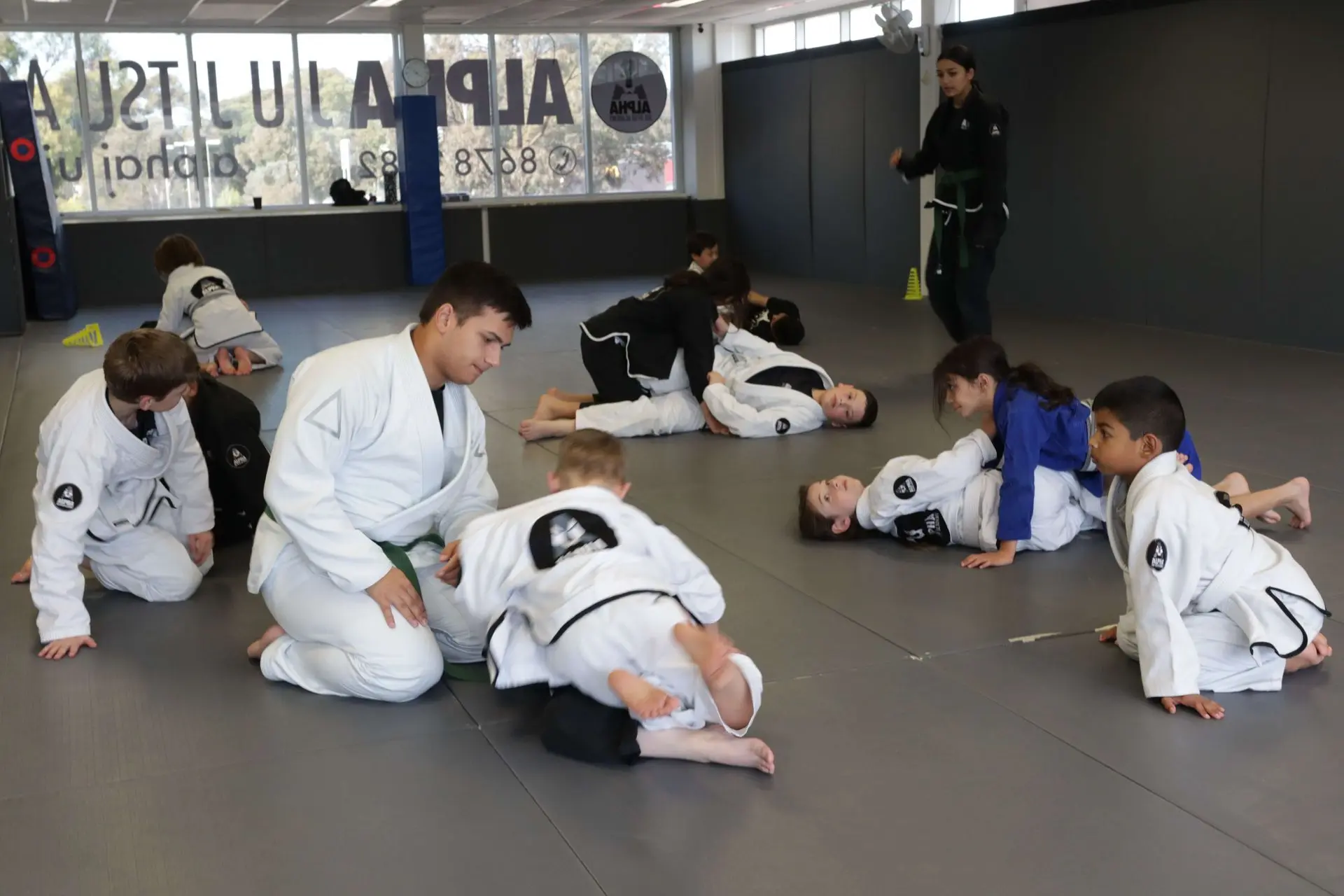 A group of young students is practicing Brazilian Jiu-Jitsu at Alpha Jiu-Jitsu Academy. In the foreground, a student in a white gi with a green belt is assisting another child who is performing a grappling maneuver. Nearby, children in white gis practice techniques while an instructor in black gi observes and provides guidance. A few students in blue and black gis are engaged in practice, rolling on the mat. The academy's logo can be seen on the walls, with other students participating actively in the session.