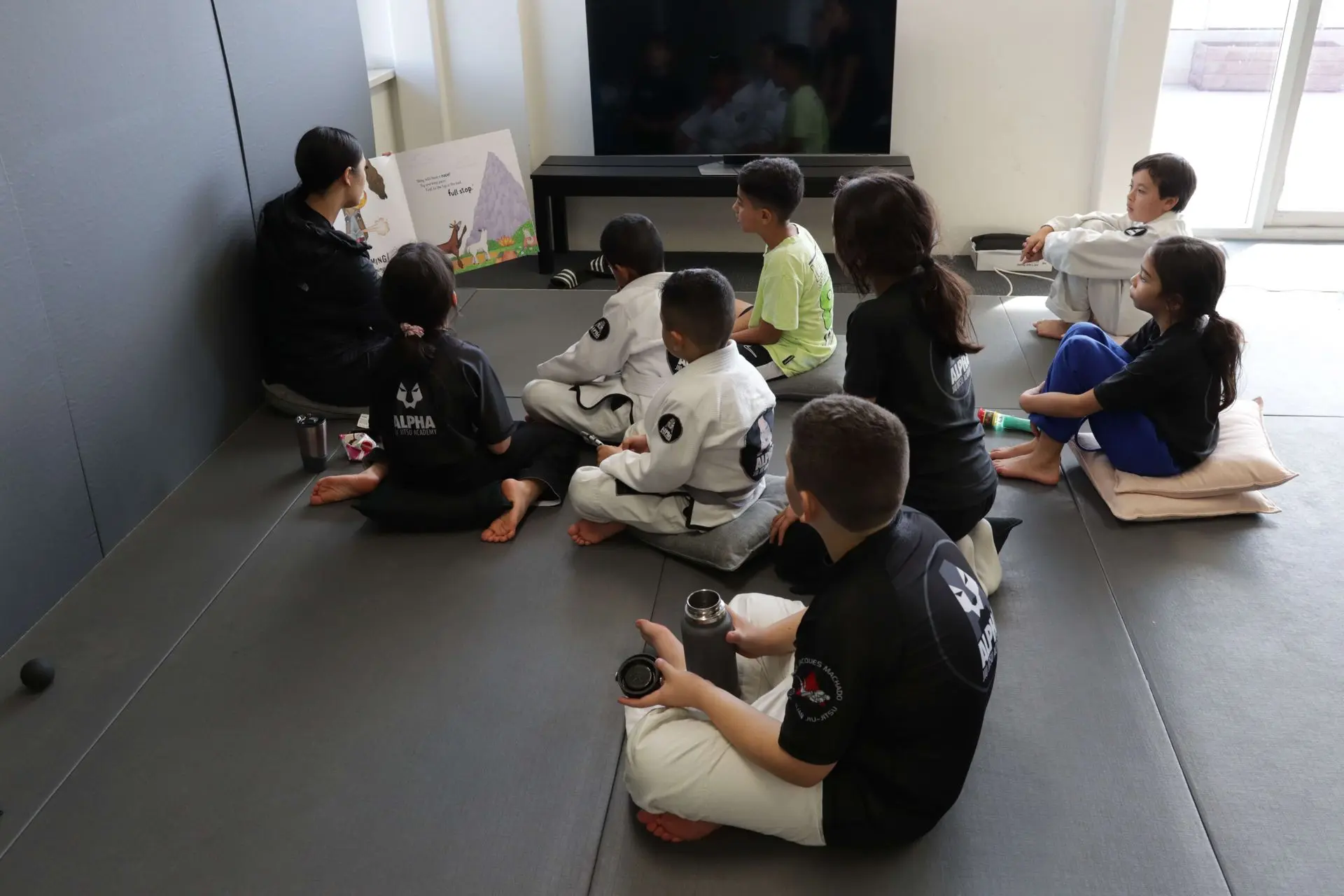 A group of children are seated on the floor, attentively listening to a story being read by an instructor at Alpha Jiu-Jitsu Academy. Some children are wearing their black Alpha Jiu-Jitsu shirts, while others are in gi uniforms. The instructor holds an illustrated children's book showing a colorful mountain scene, while the children are seated with their legs crossed or on pillows, focusing on the story. In the background, a TV and natural light from the windows add a bright atmosphere to the room.