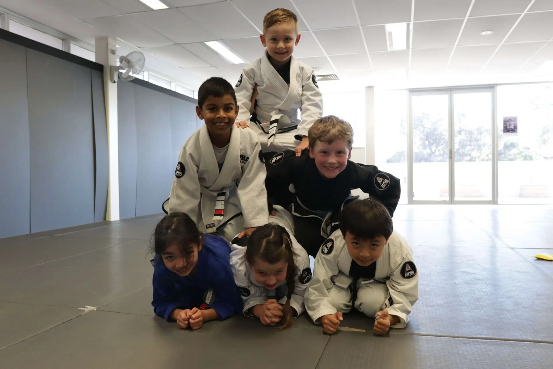 A group of kids, wearing Brazilian Jiu-Jitsu gi, are having fun by forming a pyramid on the mats of the Alpha Jiu-Jitsu Academy. The children are smiling and laughing, with some in white gis and one in a blue gi. The room has bright natural light coming from the windows in the background, highlighting the playful atmosphere and camaraderie among the group.