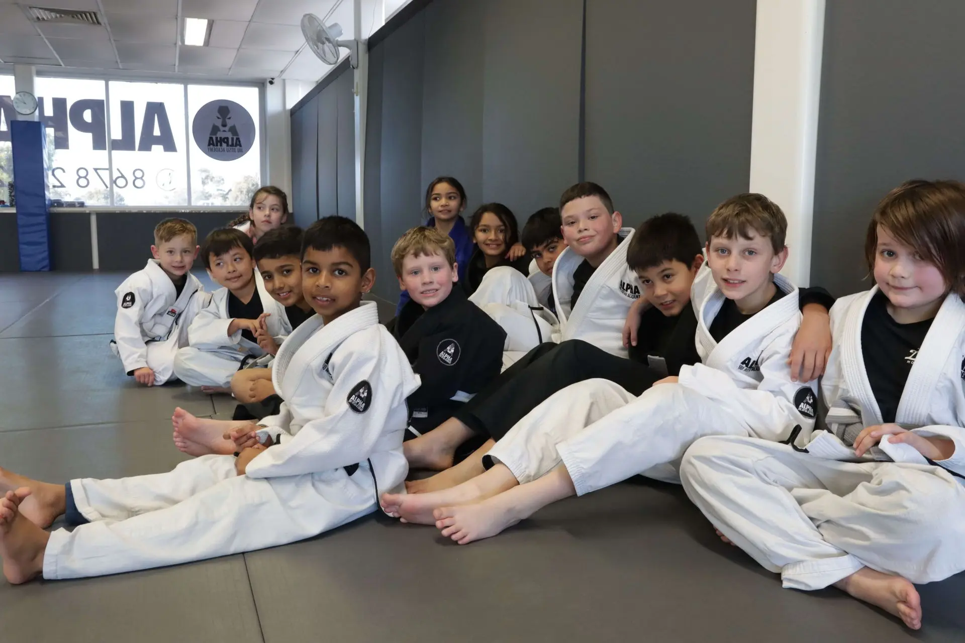 A group of young students sits together on the mat at Alpha Jiu-Jitsu Academy, smiling for the camera. They are dressed in gi uniforms, with a mix of white and black gis. The children are casually seated in a relaxed position, with some having their arms around each other, and others sitting with their legs crossed. In the background, a few other children are practicing their moves, and the academy’s logo is visible through the window on the wall. The atmosphere is cheerful and friendly.