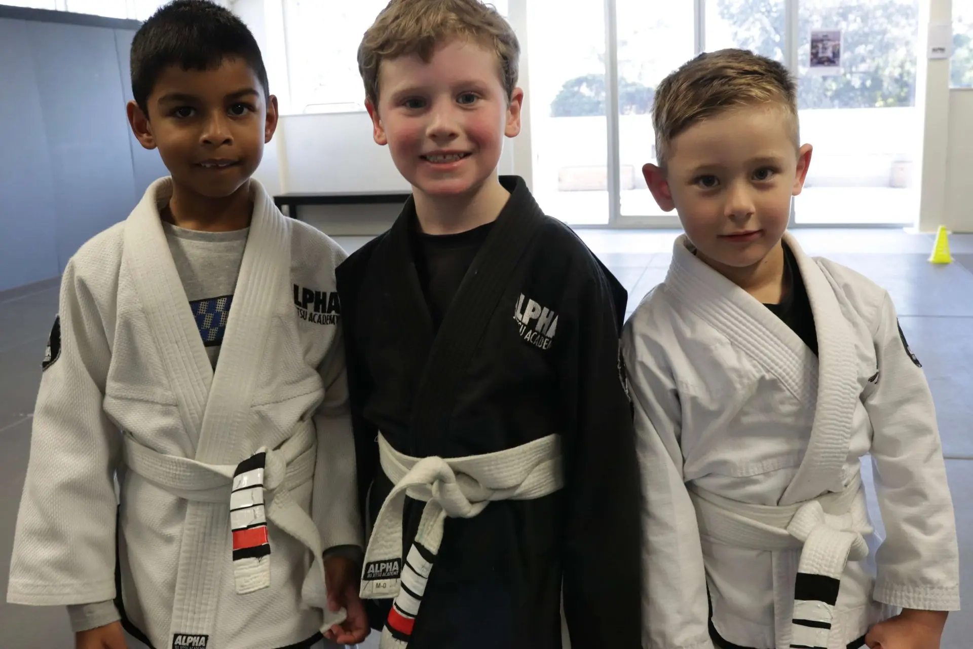 Three young children are standing side by side at Alpha Jiu-Jitsu Academy. The boy on the left is wearing a white gi with the academy's logo, the boy in the middle is wearing a black gi with a white belt, and the boy on the right is wearing a white gi with a white belt. They are smiling and looking at the camera, with a background of the academy’s training space.