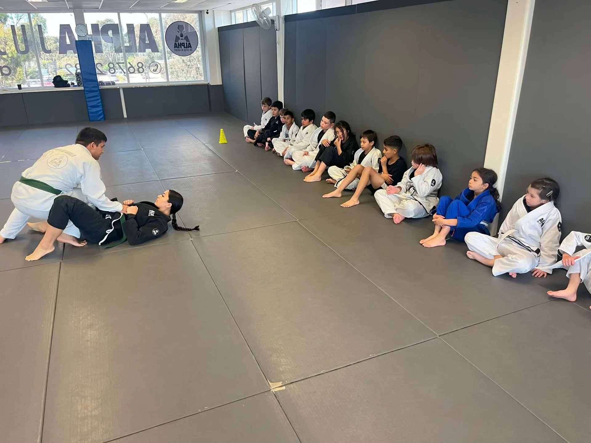 A group of children at Alpha Jiu-Jitsu Academy are either training or observing during class. In the foreground, one child in a green belt is practicing a technique on the mat with a partner. Several children sit along the wall in the background, watching attentively while others wait for their turn. The children in the sitting line are dressed in a mix of black, blue, and white gi, with a yellow cone placed on the mat to mark the training area. The academy’s branding is visible in the background.