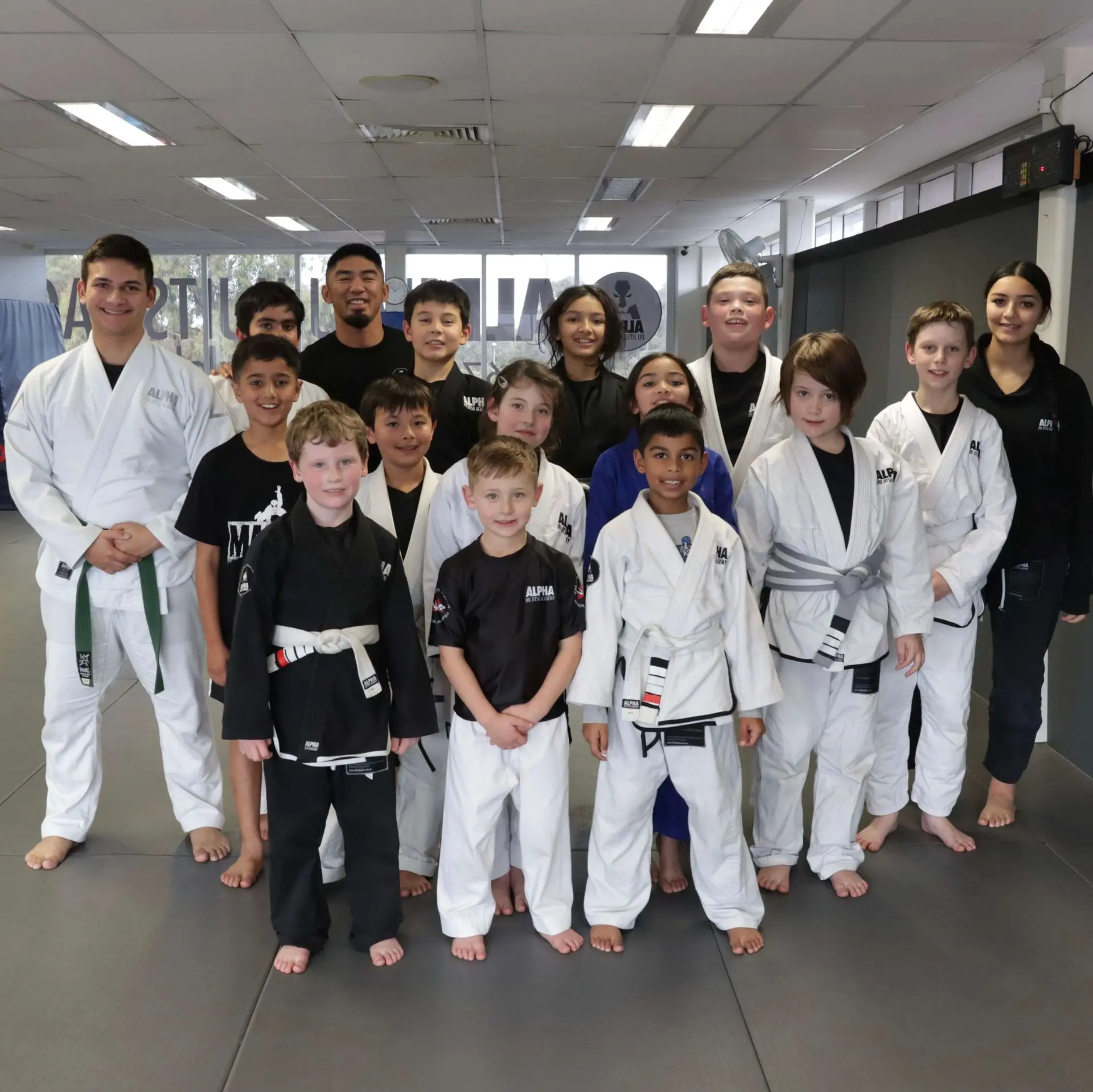 A group of smiling kids and coaches from Alpha Jiu-Jitsu Academy pose together for a photo after their class. The children, some in gi uniforms and others in athletic gear, are of various ages and belt ranks, showcasing the diversity and camaraderie within the academy. The coaches, standing at the front with the students, share a proud moment with the kids. The training mats and the academy's branding can be seen in the background, highlighting the positive and supportive environment.