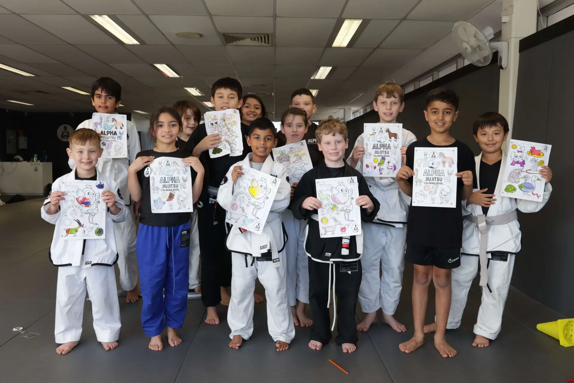 A group of happy children, dressed in Brazilian Jiu-Jitsu gis and casual clothing, proudly display their completed coloring sheets featuring Alpha Jiu-Jitsu branding. The children are standing on the mats of the Alpha Jiu-Jitsu Academy, with colorful images of characters and animals on their sheets. The room is bright with ample natural light coming through the windows, creating a warm and cheerful atmosphere. Some of the kids are in white gis, while others wear black or blue gi pants, and they all smile while holding up their artwork.