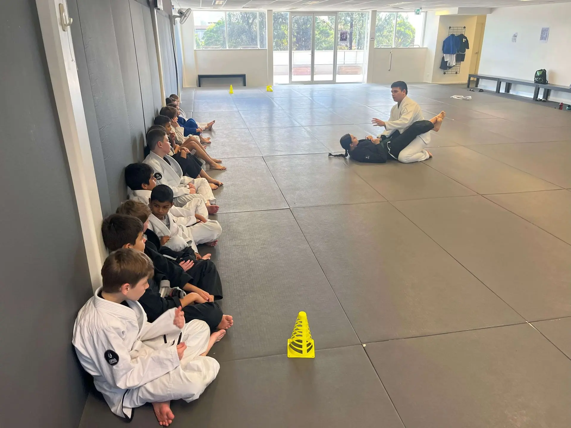 A group of children are sitting on the side of the mat, attentively watching an instructor demonstrate a technique with a student in the foreground. The instructor is showing a Brazilian Jiu-Jitsu move on a student, who is wearing a gi. The kids, wearing a mix of gi uniforms, are seated along the wall in an organized manner, with yellow cones marking sections of the mat. The academy's training space is well-lit and spacious, with neatly arranged mats and a clean environment.