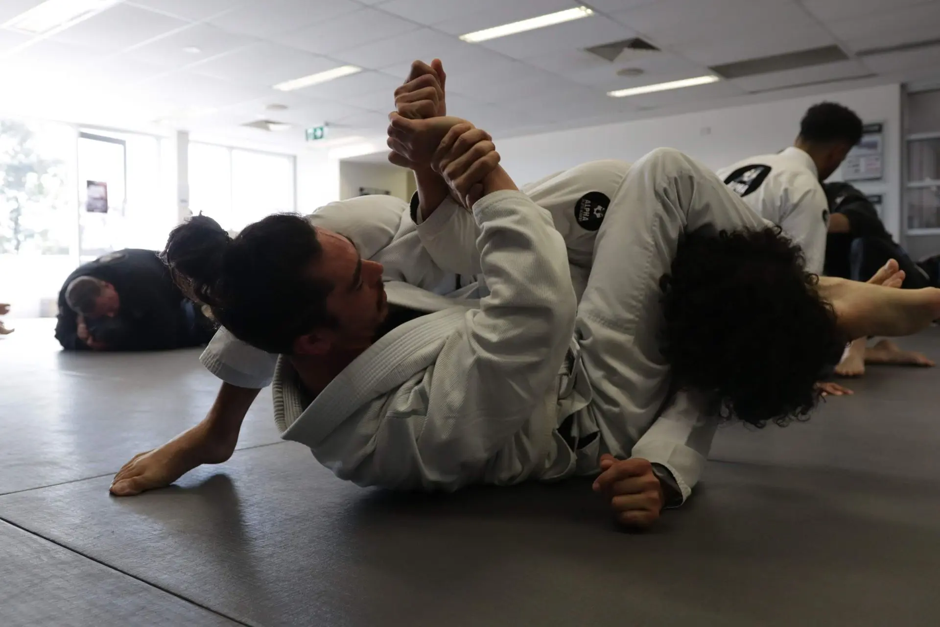 Two practitioners of Brazilian Jiu-Jitsu are engaged in a grappling session, showcasing submission techniques. One practitioner is on top in a controlling position, securing an arm hold while the other tries to defend and escape. Both are wearing gi uniforms, with Alpha Jiu Jitsu Academy patches visible. The background features other students training, with a focus on technique practice. The well-lit room creates a dynamic and focused training environment, highlighting the intensity and concentration of the grapplers.