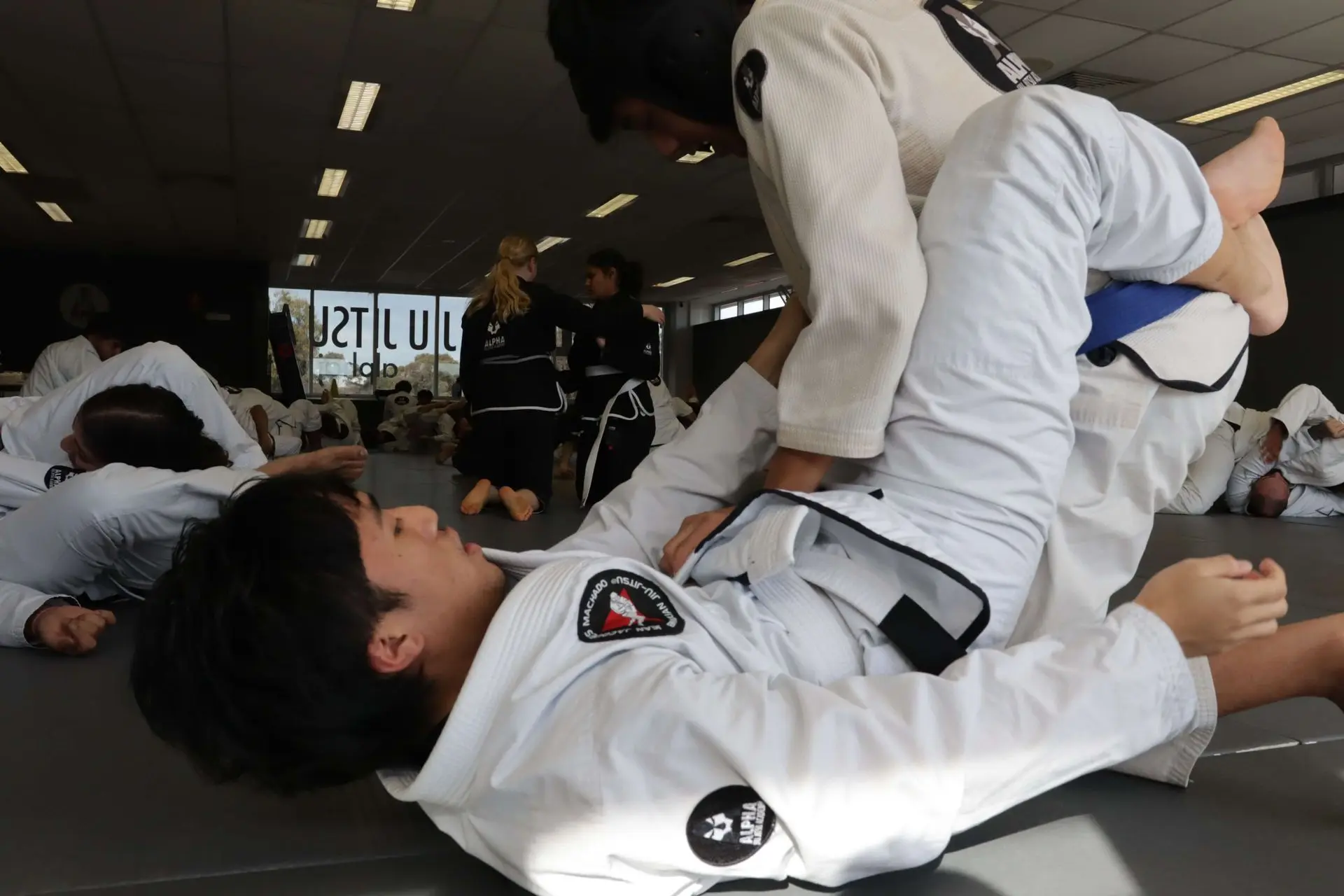 Two students practicing a jiu-jitsu guard technique during a training session at Alpha Jiu Jitsu Academy. The student on the bottom is executing a guard maneuver, while the student on top is maintaining a position, wearing a protective headgear. Other students are visible training in the background, enhancing the atmosphere of focused learning and practice.