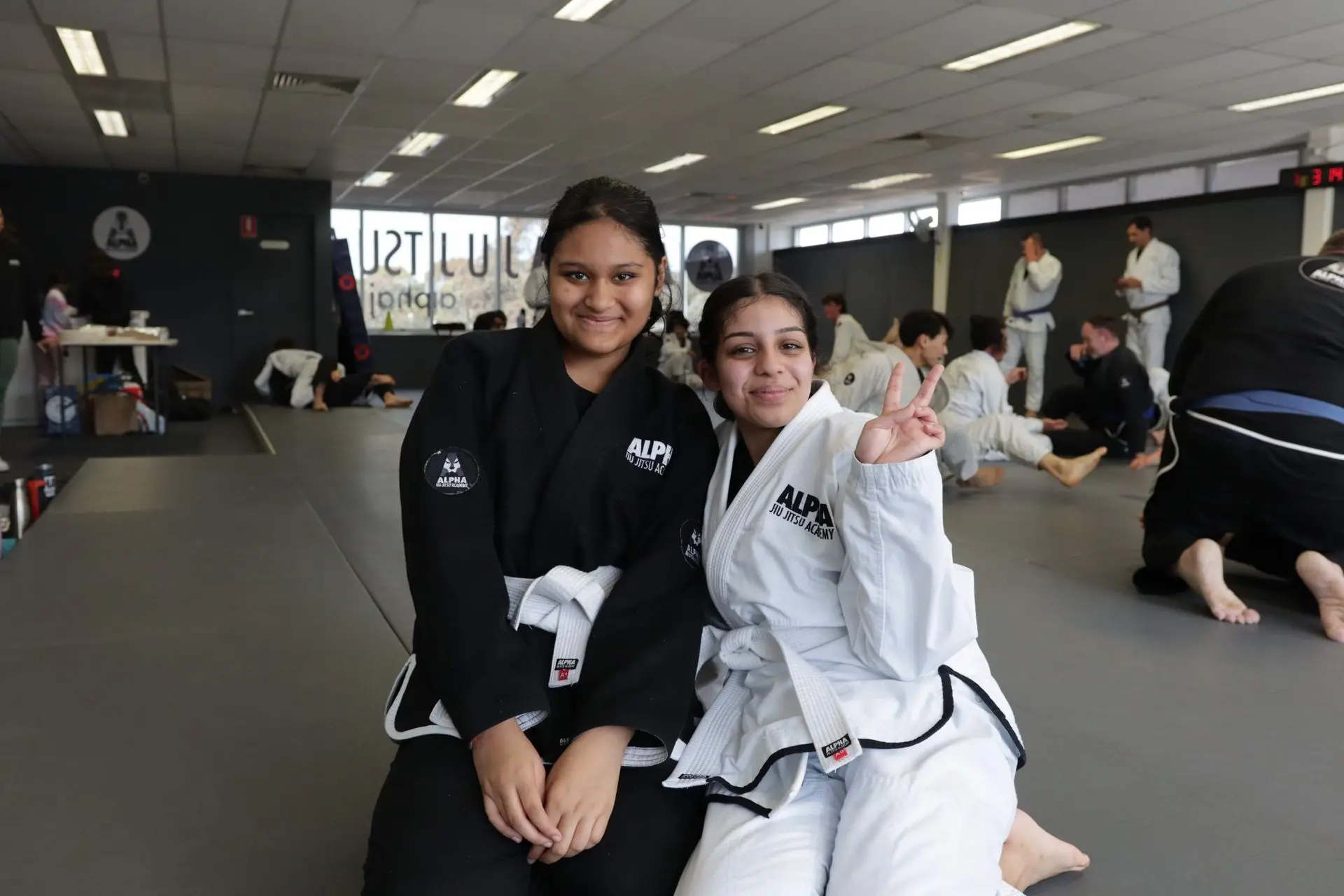 Two young women, one wearing a black gi and the other a white gi, sit together on the training mat at Alpha Jiu-Jitsu Academy. Both are smiling, with the woman in the white gi giving a peace sign. The background shows other students practicing Brazilian Jiu-Jitsu on the mat, highlighting an active and positive training environment. The academy's branding is visible on their gi patches, and the space is well-lit, offering a friendly and welcoming atmosphere for learners.