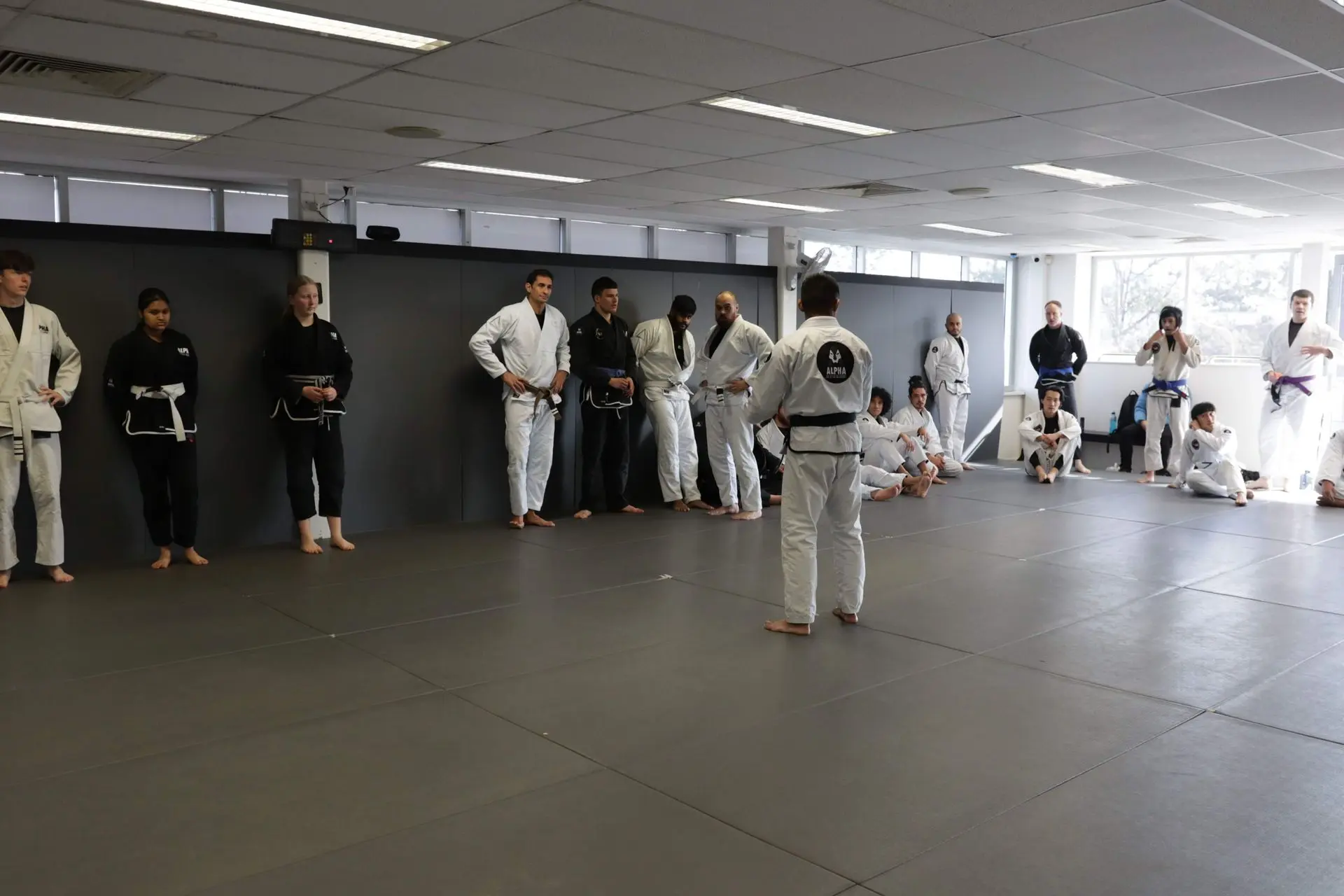 Students and instructors at Alpha Jiu Jitsu Academy are engaged in a lesson. A senior instructor is speaking to the group, with some students standing and others sitting on the floor in a relaxed yet attentive posture. The participants are in various gi uniforms, with belts indicating their rank, such as white, blue, and black belts. The room is well-lit and spacious, with large windows that allow natural light to flood the training area. The class appears focused on learning and practicing techniques, with a few students actively taking notes or preparing for their turn.