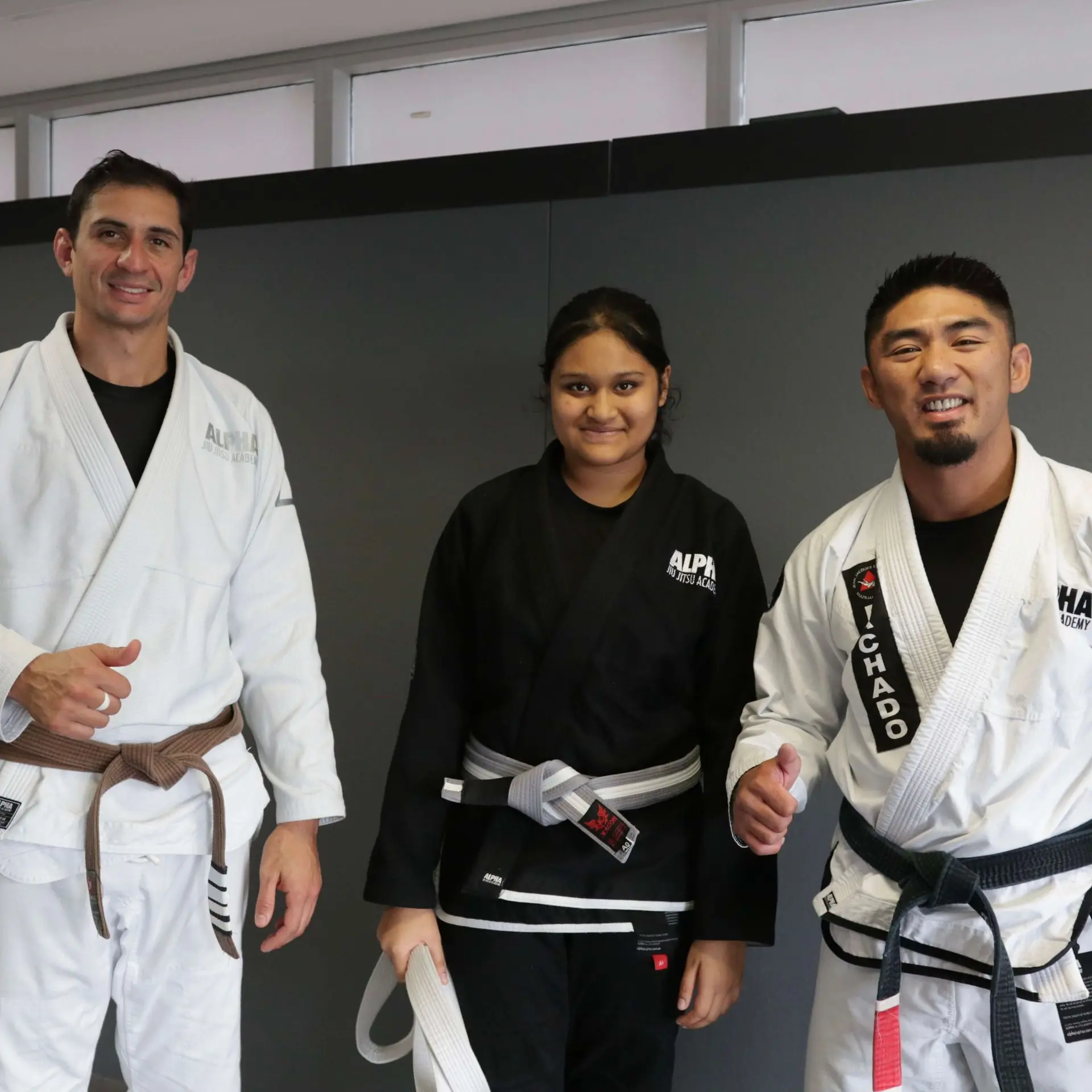 Three individuals at Alpha Jiu Jitsu Academy stand proudly, with the middle student wearing a black gi and holding a new belt. Flanked by two instructors in white gis, all three smile warmly, celebrating the student's milestone achievement in martial arts training. The atmosphere reflects camaraderie, support, and growth within the academy's community.