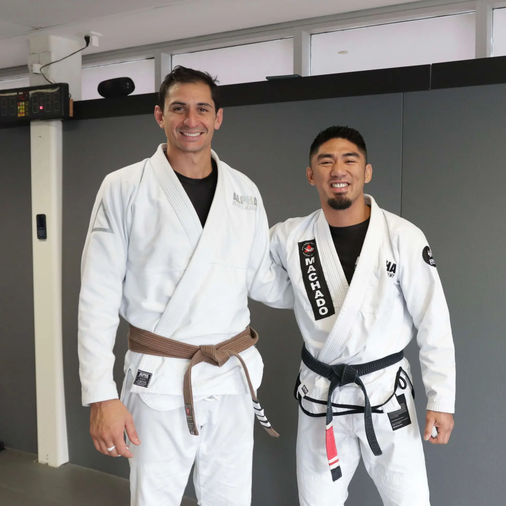 Two instructors from Alpha Jiu Jitsu Academy, one wearing a brown belt and the other a black belt embroidered with "Machado," stand side by side, smiling. The image highlights the welcoming and experienced teaching environment of the academy.