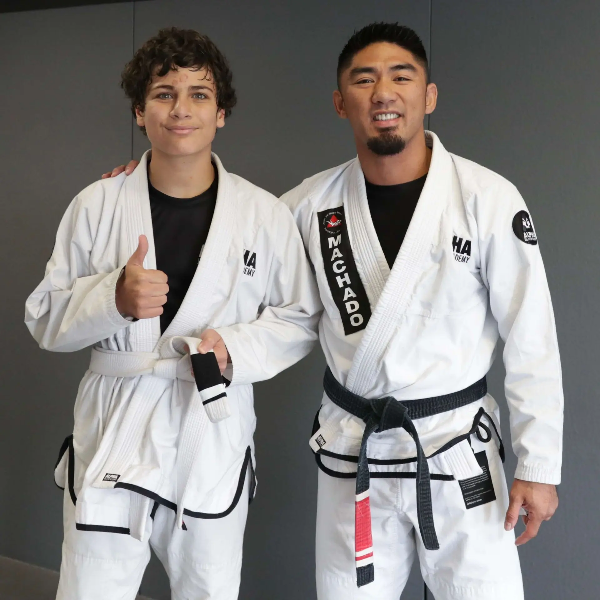 A student wearing a white belt poses with a thumbs-up alongside a black belt instructor at Alpha Jiu Jitsu Academy. The instructor's belt is embroidered with the name "Machado," showcasing their expertise. This moment captures the celebration of progress and encouragement within the academy's training environment.