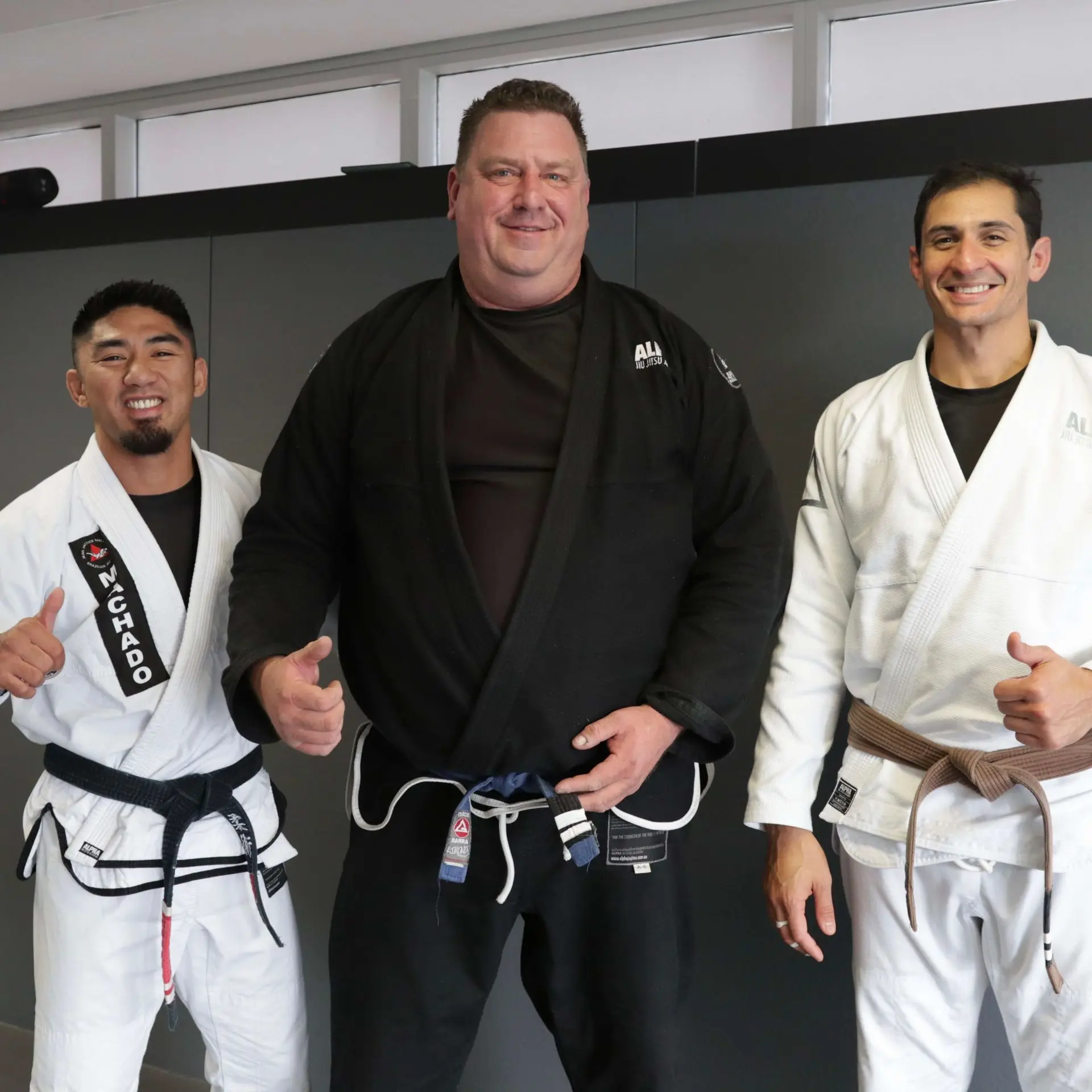 A photo of three individuals standing together at Alpha Jiu-Jitsu Academy. On the left, a Brazilian Jiu-Jitsu instructor wearing a white gi and black belt gives a thumbs up. In the middle, a student is wearing a black gi and a blue belt, smiling and also giving a thumbs up. On the right, another instructor is dressed in a white gi with a brown belt, also smiling and giving a thumbs up. They all stand in front of a dark gray wall in the academy.