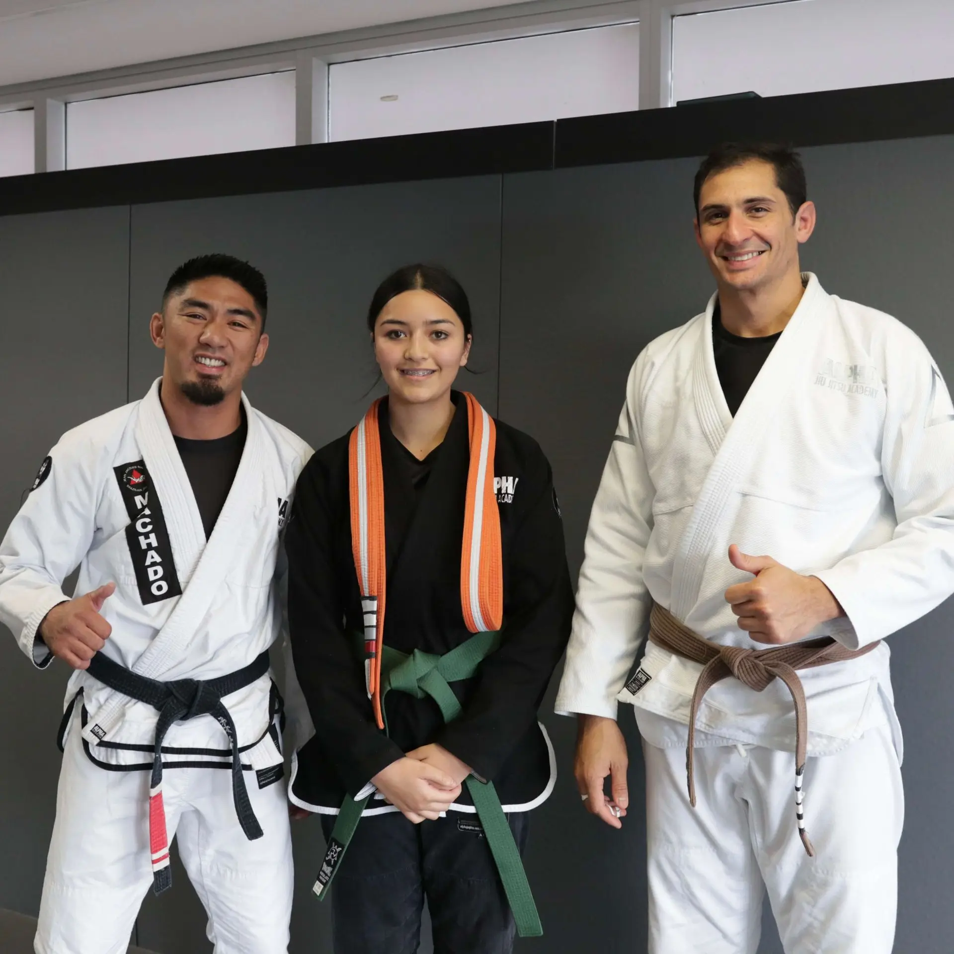 A group photo showing three people at Alpha Jiu-Jitsu Academy. On the left is a male instructor wearing a white gi and black belt, smiling and giving a thumbs up. In the middle, a female student is wearing a black gi with a green belt and two orange-striped belts over her shoulder, smiling. On the right, another male instructor is wearing a white gi with a brown belt, also smiling and giving a thumbs up. They are posing in front of a gray wall inside the academy.