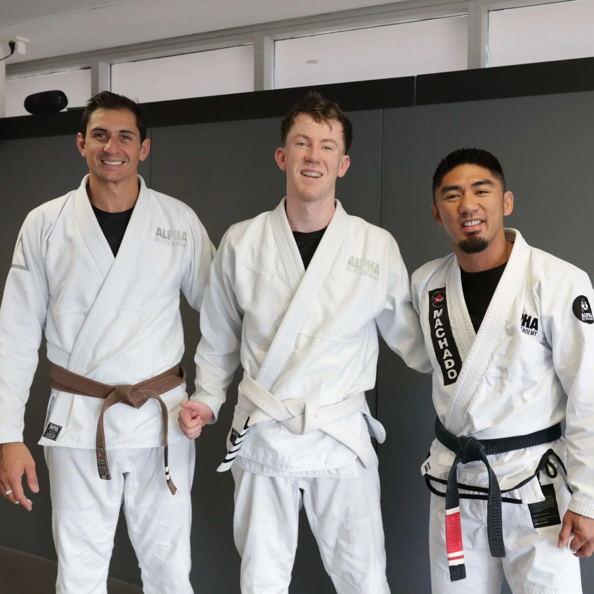 A student at Alpha Jiu Jitsu Academy stands between two instructors, all wearing white gis and smiling for the photo. The student, who has earned a new belt, is flanked by supportive instructors, signifying a proud moment of accomplishment in the academy. The image captures the spirit of mentorship, encouragement, and growth within the Alpha Jiu Jitsu Academy community.