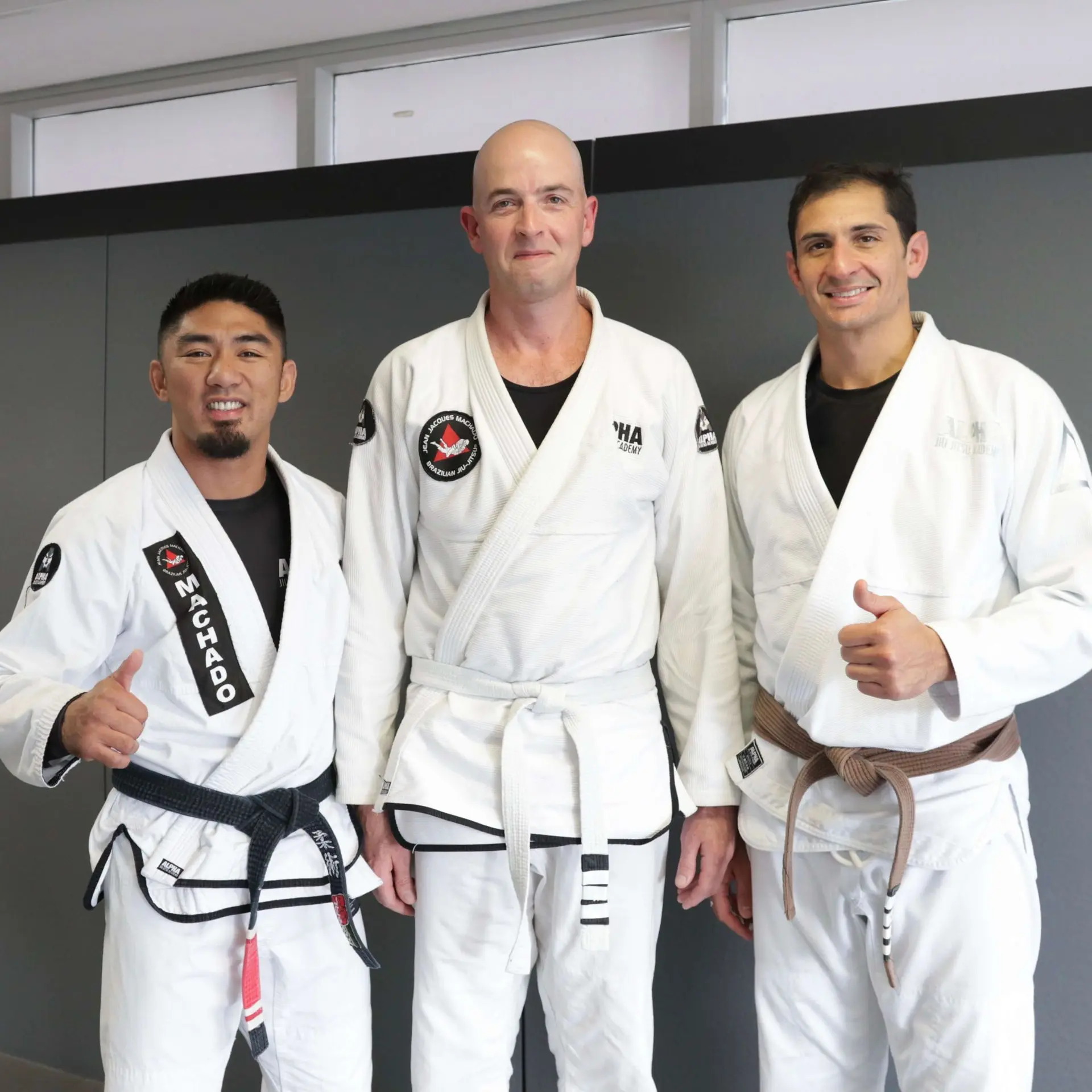 Three members of Alpha Jiu Jitsu Academy standing together in white gi uniforms, one with a black belt featuring "Machado," one with a white belt, and one with a brown belt, smiling and giving thumbs up, representing camaraderie and expertise at the academy.