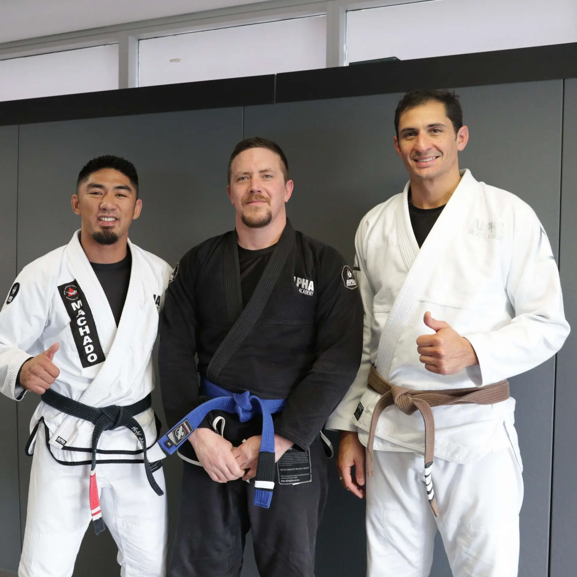 Three members of Alpha Jiu Jitsu Academy posing together, with the member in the center wearing a blue belt and black gi, flanked by two instructors in white gis with brown and black belts, giving a thumbs-up and smile, symbolizing teamwork and dedication at Alpha Jiu Jitsu Academy.