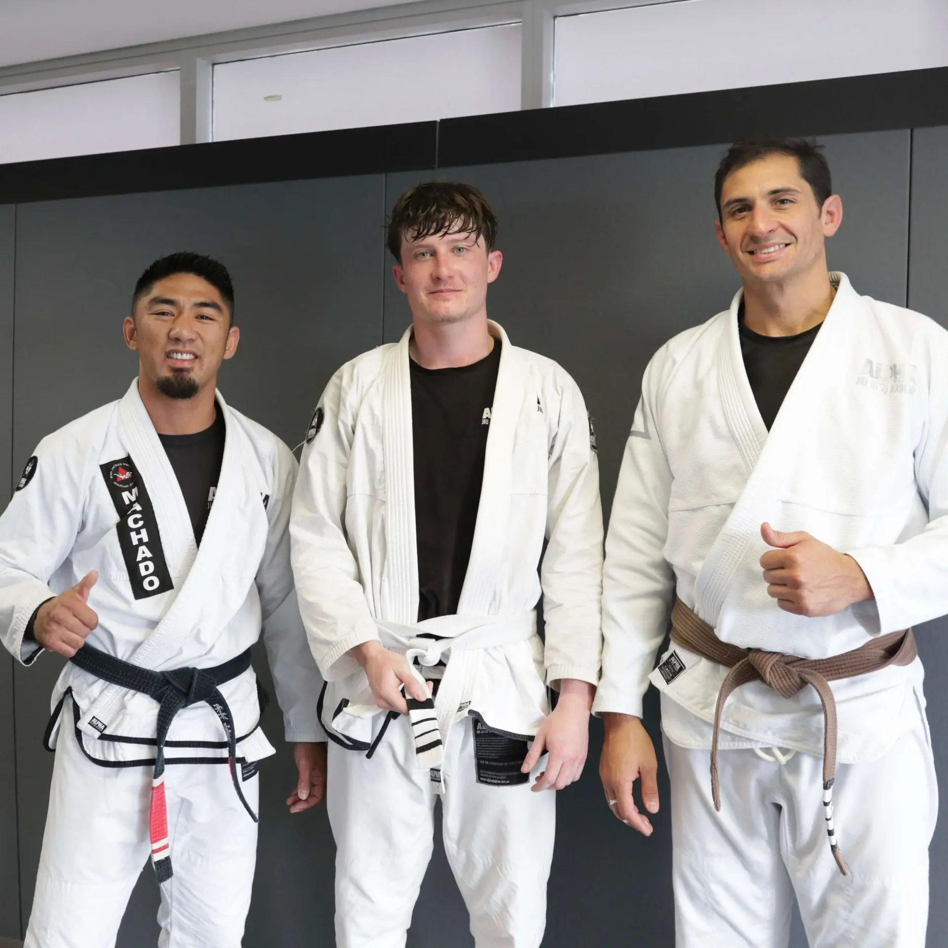 A proud student, fresh from his hard-earned white belt promotion, stands with two instructors at Alpha Jiu Jitsu Academy. The instructor on the left, wearing a black belt, and the instructor on the right, wearing a brown belt, both smile and give thumbs up in celebration. The student holds his new white belt and looks excited after reaching this milestone. The clean, modern background highlights the academy's professional setting.