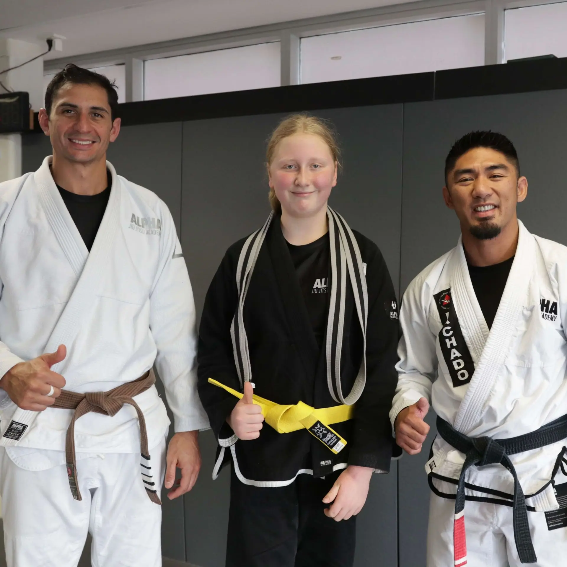 A student proudly stands between two instructors at Alpha Jiu Jitsu Academy, all smiling and giving a thumbs-up. The student is wearing a black gi with a newly earned yellow belt and a striped belt draped over their shoulders. The instructors, wearing white gis, show their support and pride for the student's achievement, reflecting the positive, encouraging environment at the academy.