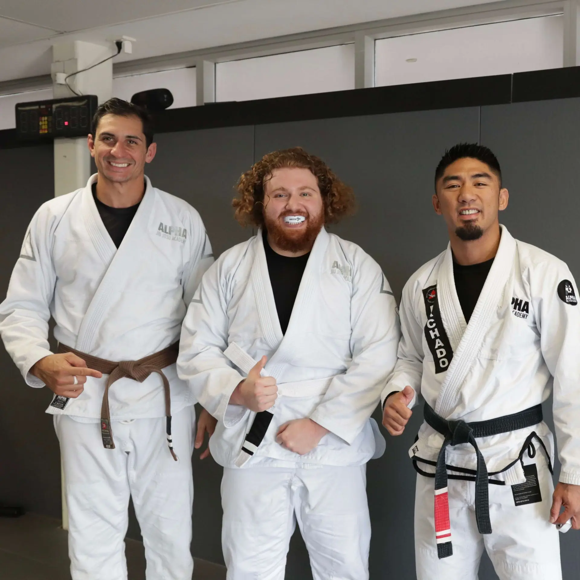 The joyful moment of a belt promotion is captured at Alpha Jiu Jitsu Academy as a student, wearing a white belt and a mouthguard, poses alongside two instructors. The instructor on the left is wearing a brown belt, while the instructor on the right sports a black belt. All three participants give thumbs up, celebrating the student’s progress in the academy. The setting is professional, highlighting the academy's modern and clean environment.