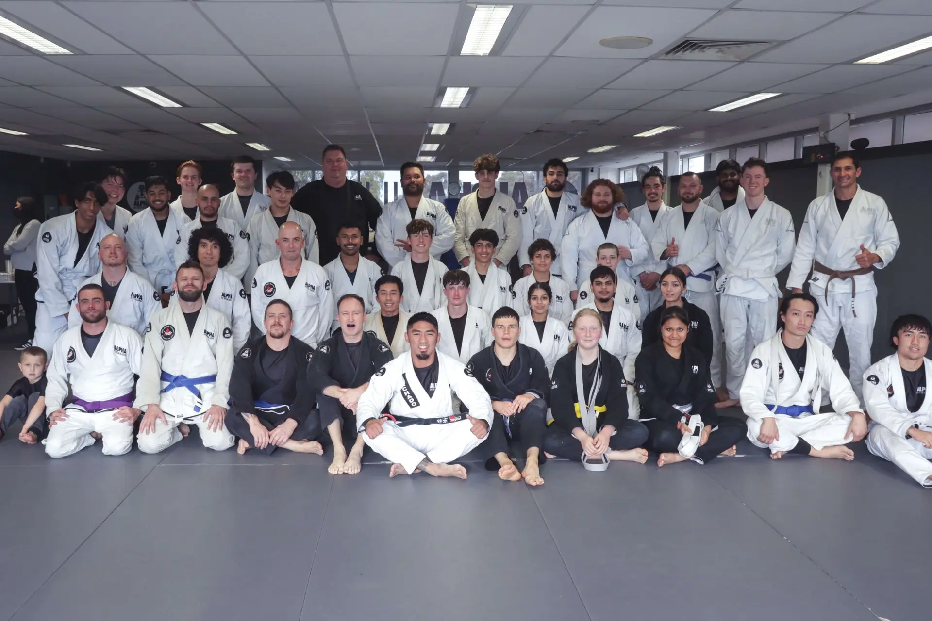 A large group of students and instructors at Alpha Jiu Jitsu Academy gathered for a class photo. Everyone is wearing their gis, showcasing a range of belt colors from white to black, symbolizing the diverse skill levels and the welcoming training environment. The group is smiling and sitting and standing together on the mats, representing camaraderie, community spirit, and dedication to Brazilian Jiu-Jitsu practice.