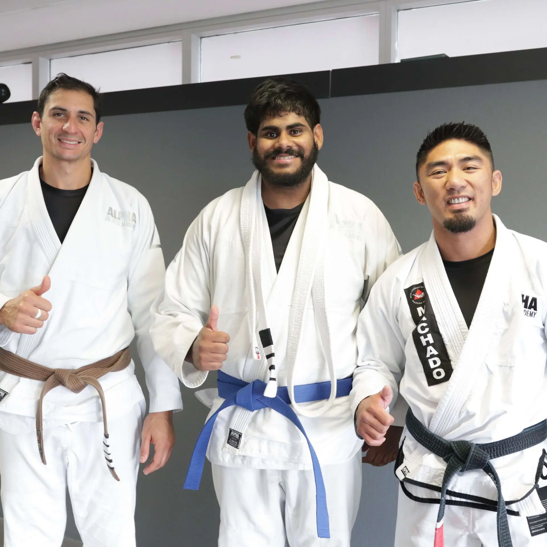 A proud student receives his blue belt promotion at Alpha Jiu Jitsu Academy. The student, smiling widely, is flanked by two instructors, each giving a thumbs-up in celebration. The instructor on the left wears a brown belt, and the instructor on the right wears a black belt. The student is wearing a white gi with a newly earned blue belt. The photo captures a joyful moment of achievement and camaraderie in front of a simple grey backdrop, with Alpha Jiu Jitsu Academy branding visible on their gis.