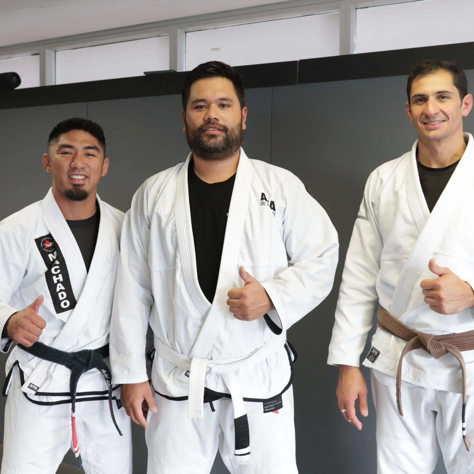 A group photo with three people standing together at Alpha Jiu-Jitsu Academy. On the left, a male instructor wearing a white gi and black belt gives a thumbs up. In the middle, a male student is wearing a white gi and black belt, standing confidently. On the right, another male instructor wearing a white gi with a brown belt is also giving a thumbs up. They are posing in front of a gray wall inside the academy.