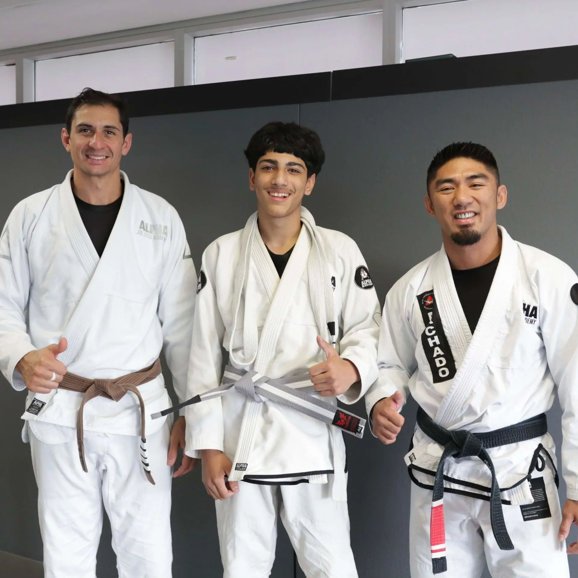 A photo of three people standing together in a martial arts studio. On the left, a male instructor in a white gi and brown belt is smiling and giving a thumbs up. In the center, a young male student is wearing a white gi with a gray belt, smiling. On the right, another male instructor in a white gi and black belt is also smiling and giving a thumbs up. The background shows a gray wall in the academy.