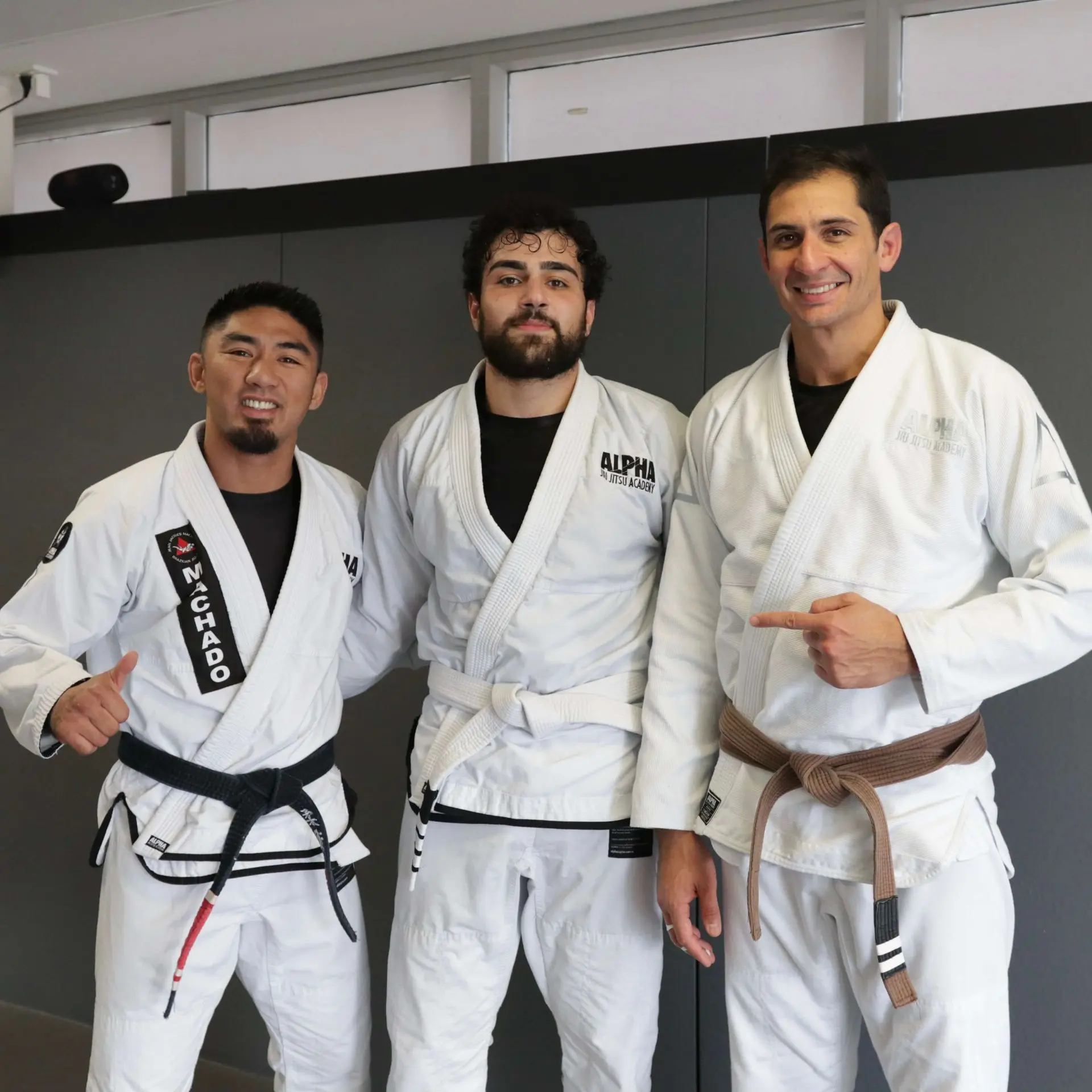 A photo of three men standing together in a martial arts studio. On the left, a male instructor in a white gi and black belt is smiling and giving a thumbs up. In the center, a male student is wearing a white gi with a white belt and a serious expression. On the right, another male instructor is pointing toward the student while wearing a white gi and brown belt, smiling. The background shows a gray wall in the academy.